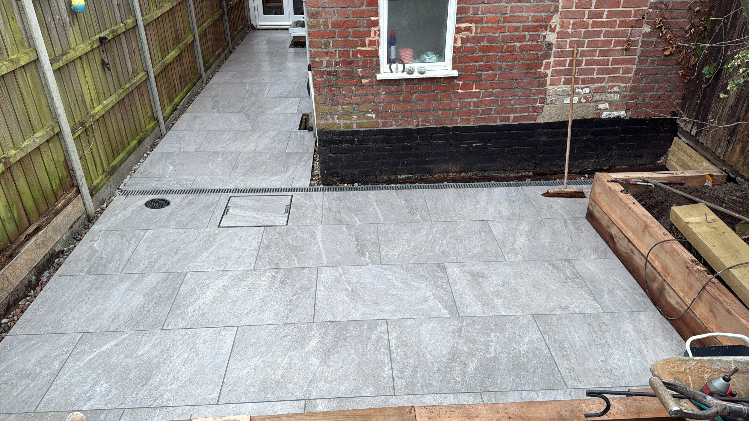 Photo of a partially completed outdoor patio area with gray tiled flooring, a brick house wall, a wooden fence, and gardening tools and construction materials around.