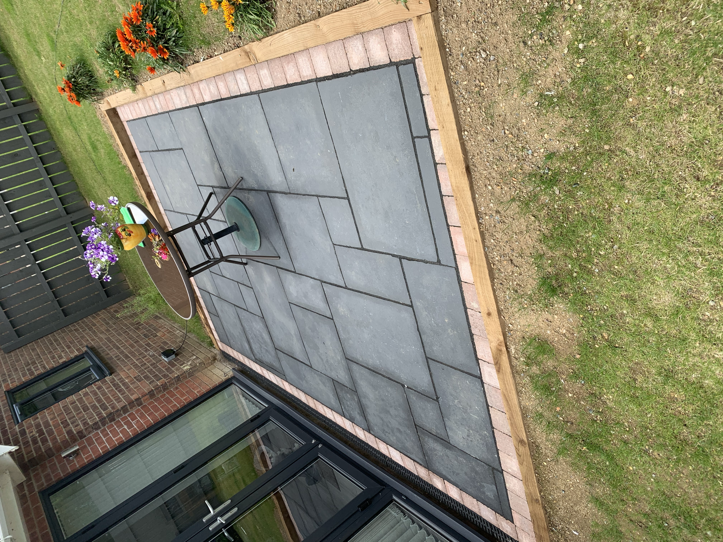 A backyard patio area with gray paving stones, bordered by a wooden frame and brick edging, adjacent to a grassy lawn with orange and purple flowers. There is a black metal outdoor table with a yellow vase of purple and white flowers on it.