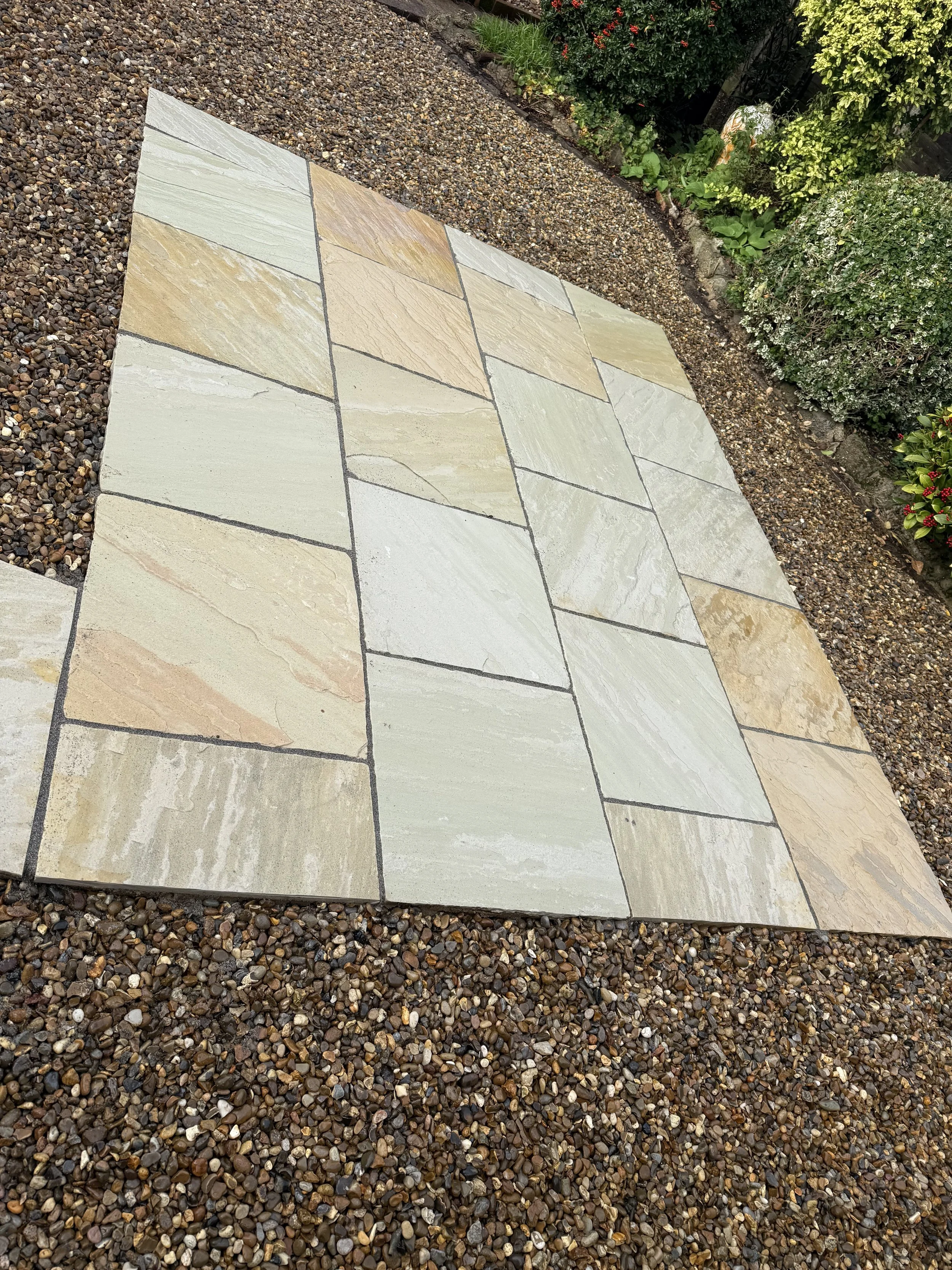 Rectangular stone tiles arranged on a gravel surface in a garden area with bushes and flowers along the edge.