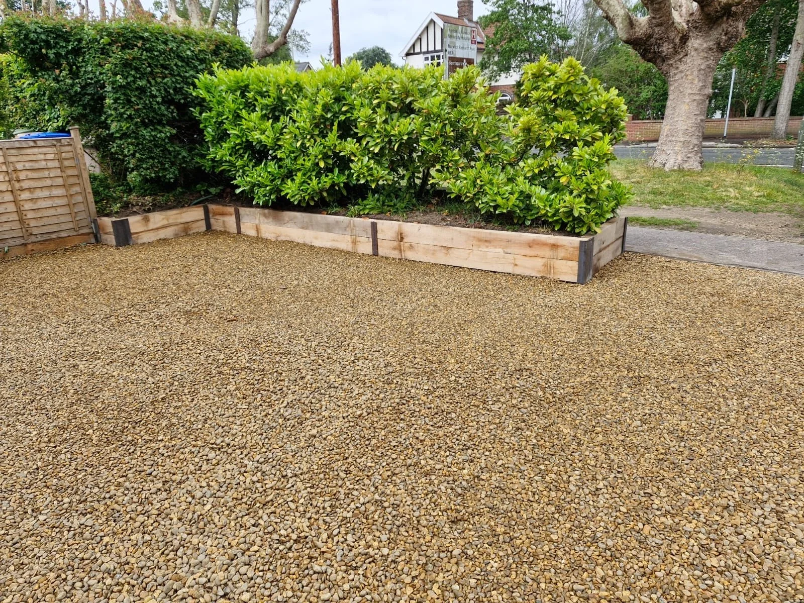 A backyard garden area with a raised wooden planter containing green bushes, a gravel yard, a wooden fence, trees, and a house in the background.