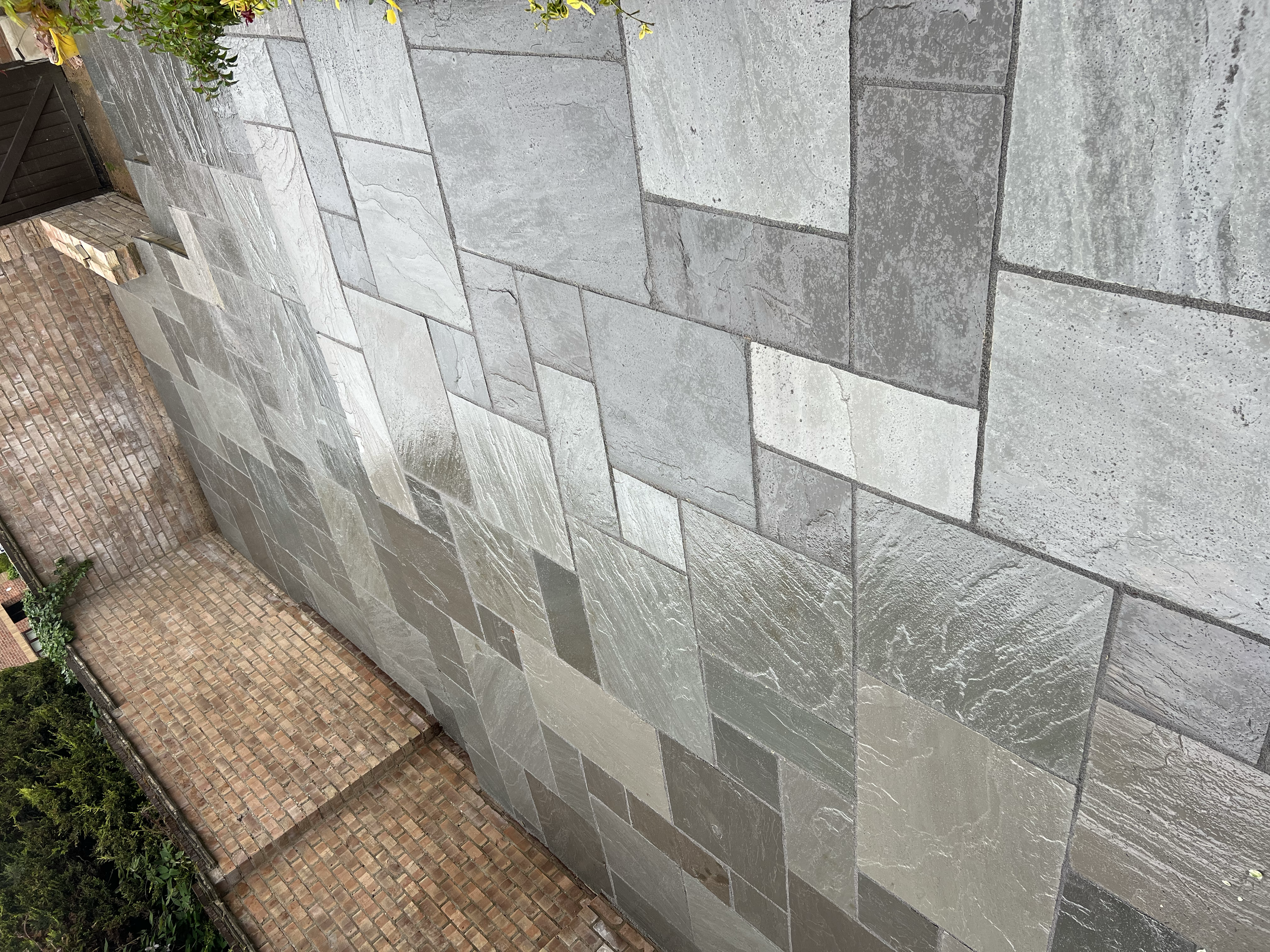 A brick wall with a dark wooden gate at the corner, next to a grey tiled patio with a leaf in the corner of the tiles.