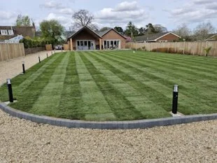 Backyard with a lush, striped lawn, a gravel pathway in the foreground, and a house with a gable roof in the background, fenced with wood panels.