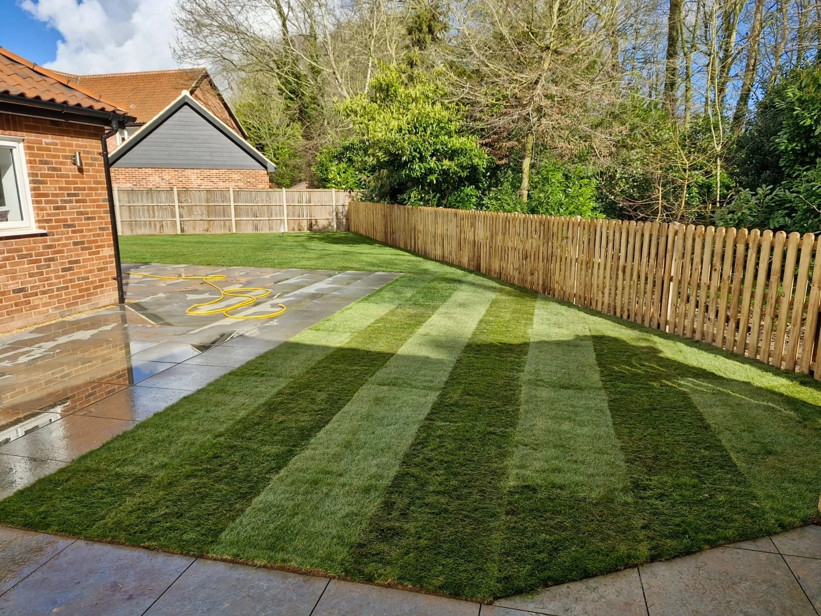 A backyard with lush green grass, a wooden fence, and a brick house. The grass has visible mowing lines, and a yellow garden hose is on wet paving stones near the house. There are trees in the background under a partly cloudy sky.