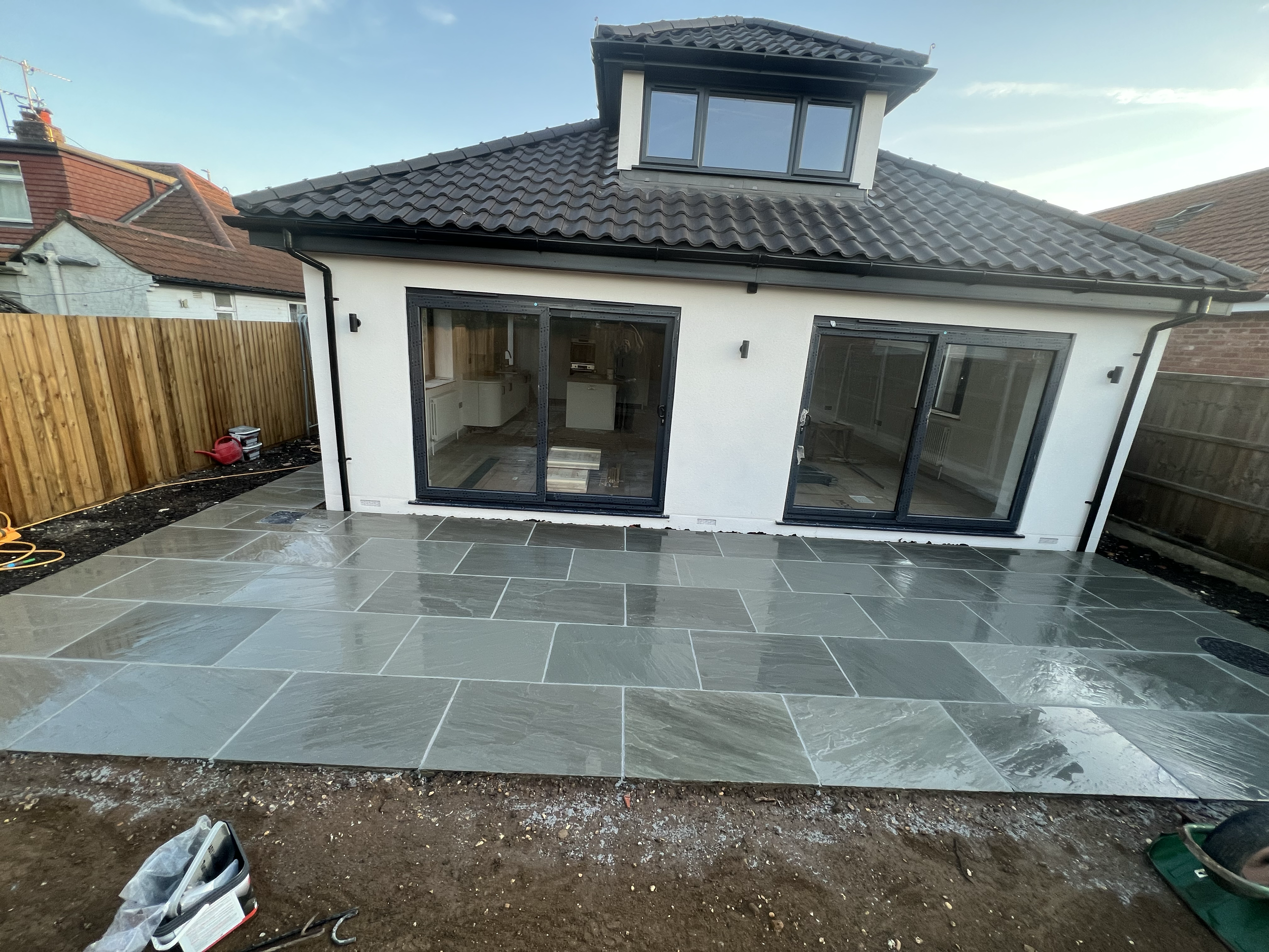 This image shows the back of a house with a new white exterior and large sliding glass doors. There is a dormer window on the roof. The area in front of the house has gray, shiny tiles, and the surrounding ground shows soil and construction tools, in