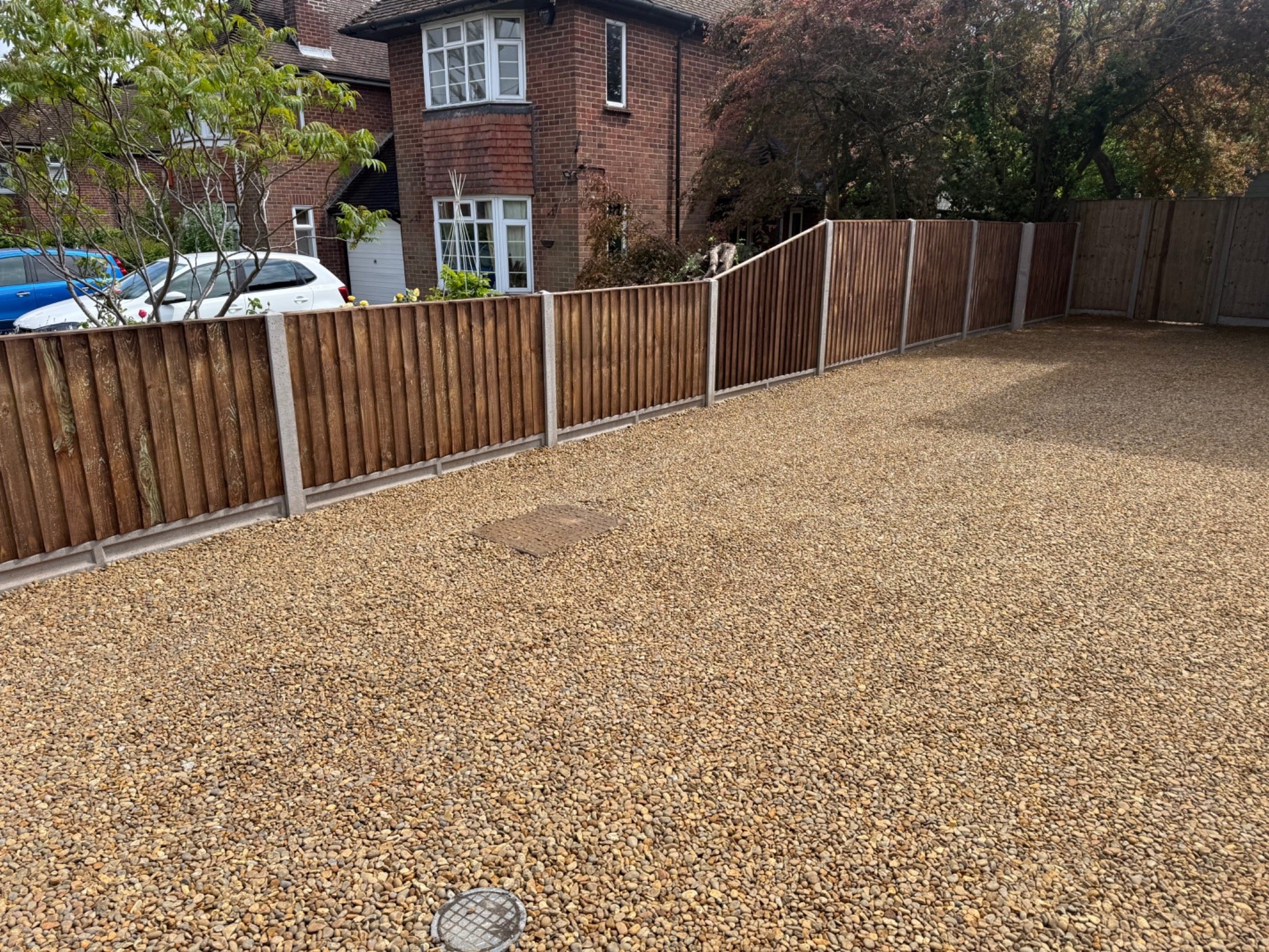 View of a gravel driveway with a wooden fence, part of a brick house, and some cars parked outside.