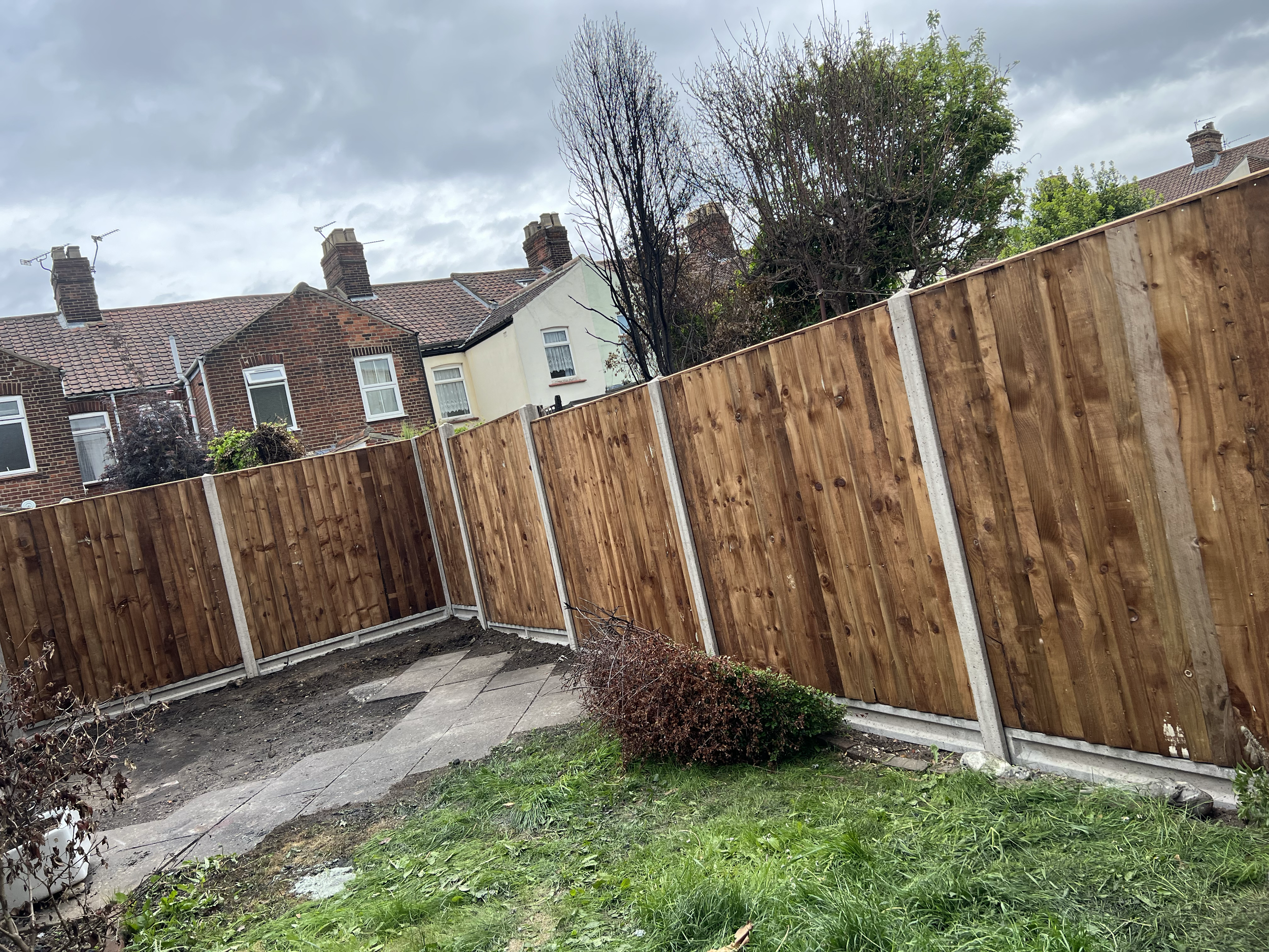 A backyard with a wooden fence, some greenery, and neighboring houses with chimneys and tile roofs under a cloudy sky.