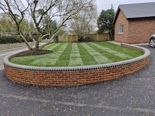 A curved brick retaining wall surrounds a small landscaped area with grass and trees, adjacent to a driveway and a house.