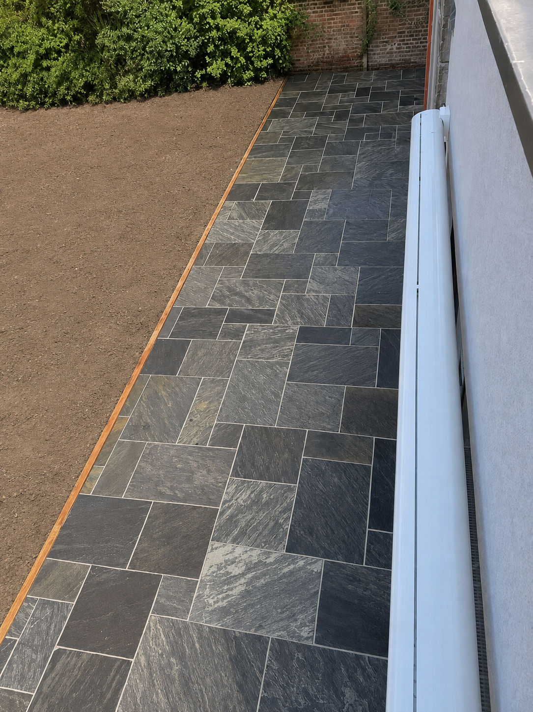 A freshly paved stone patio next to a house, bordered by a wooden strip, with a white rain gutter on the wall and green bushes in the background.