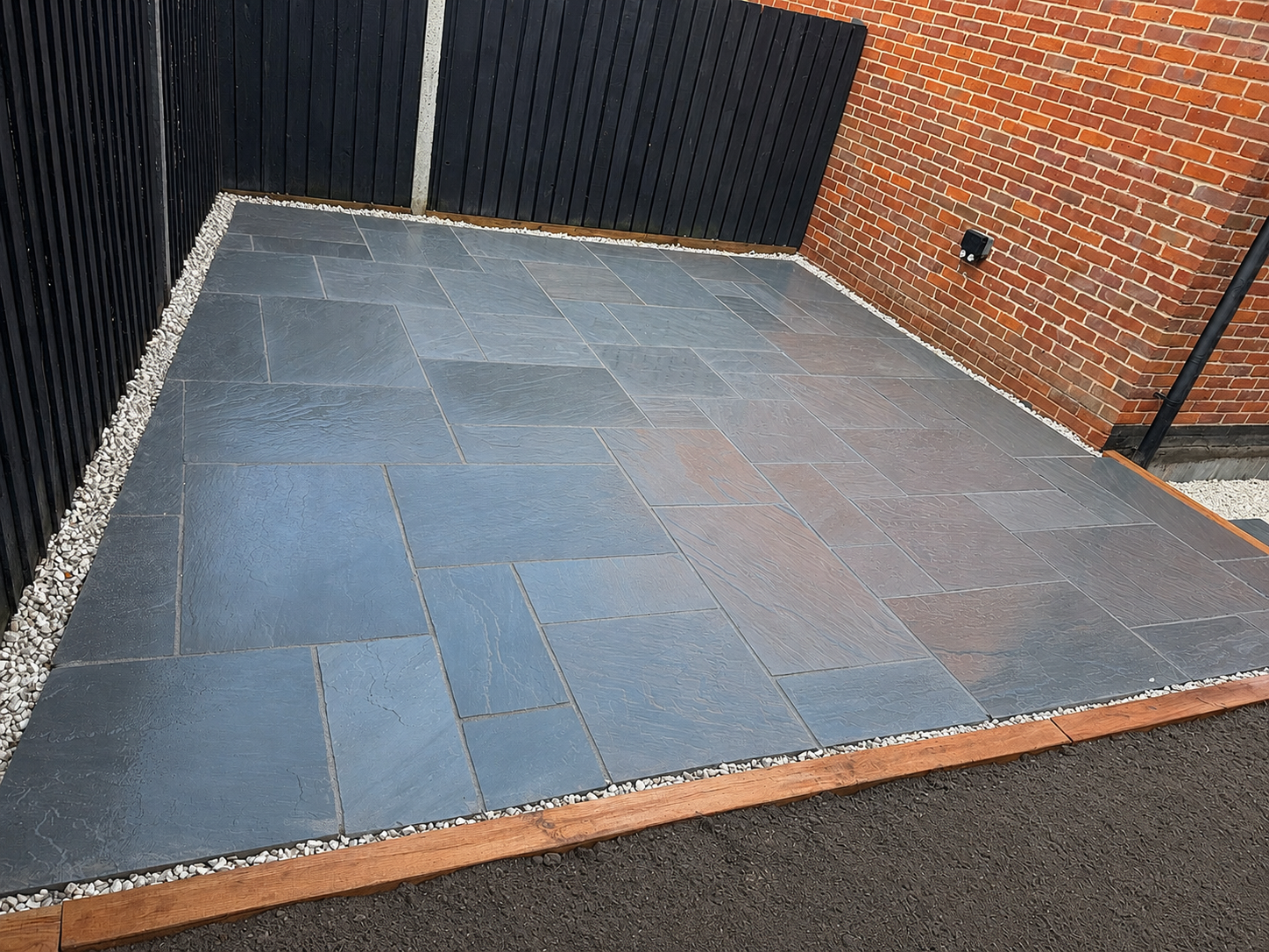 A small outdoor patio with large dark gray slate tiles, bordered by small white gravel, adjacent to a red brick wall and black wooden fence.