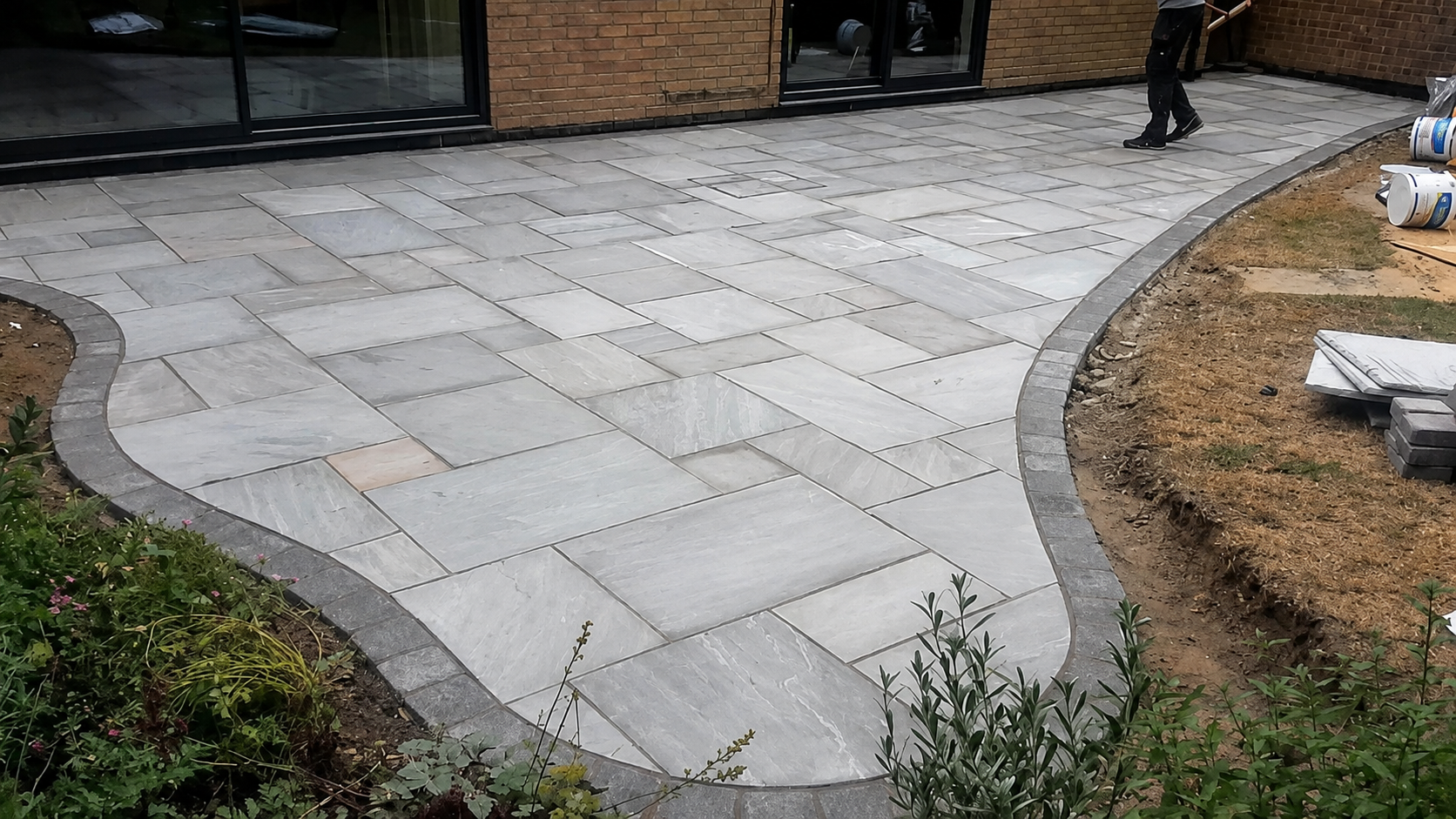 Newly installed stone patio with a curved border, adjacent to a building with glass doors and construction materials nearby, and some plantings on the side.