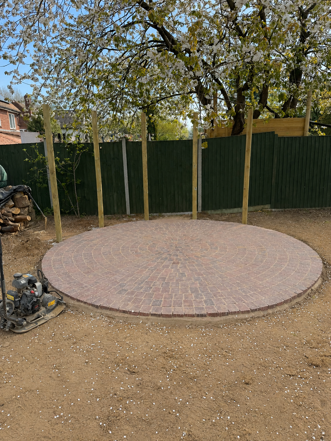 A circular brick patio under construction in a backyard, with wooden posts installed around the perimeter and a background of a green fence, blooming tree, and neighboring houses.