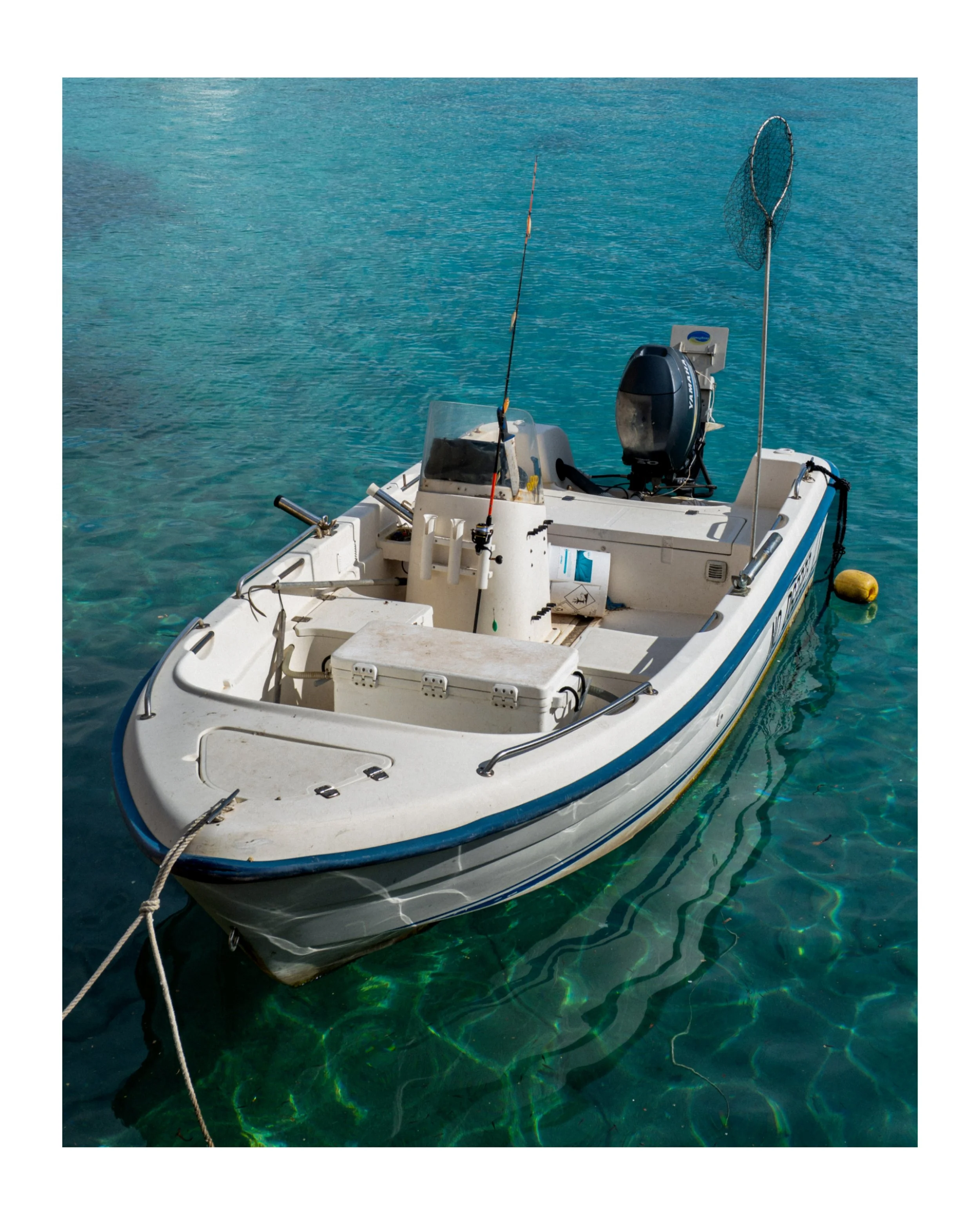 Small white boat on clear, greenish-blue water with fishing rod, kayak paddle, and small outboard motor.