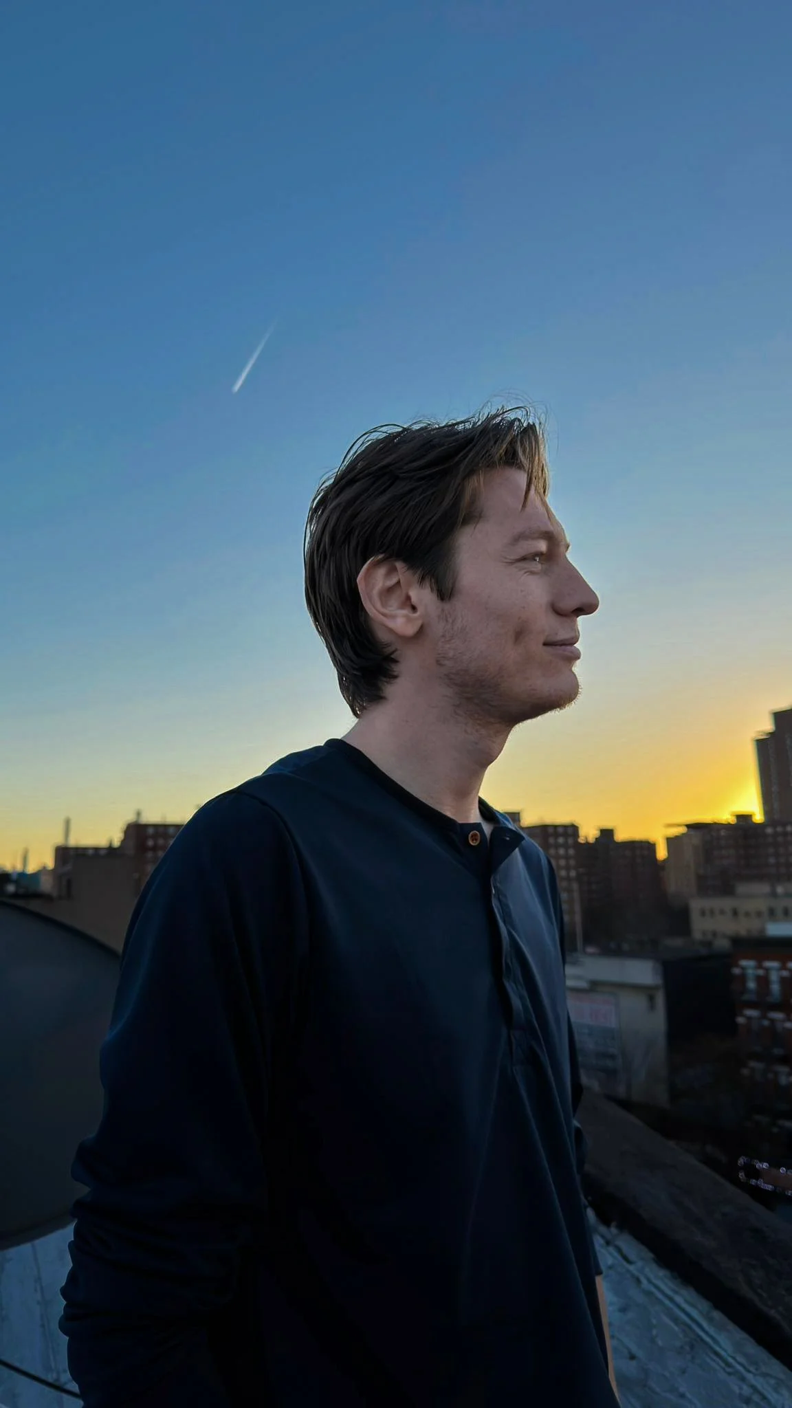 A young man with brown hair and light skin is standing on a rooftop at sunset, facing right with a city skyline in the background and a visible contrail in the sky above.