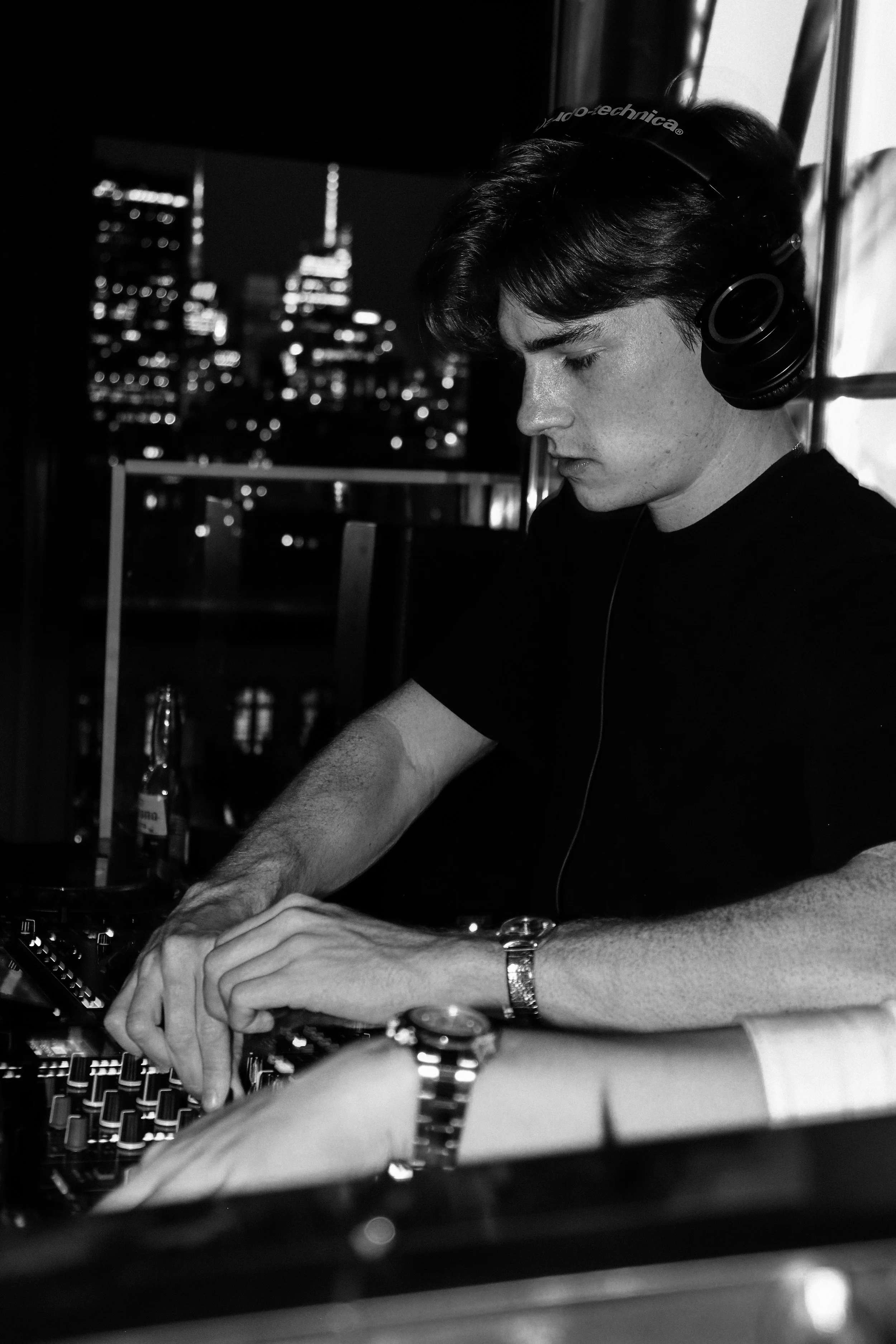 A young man with dark hair and a watch on his wrist, wearing headphones, is working on DJ equipment at a nightclub or event, with a city skyline visible through a window in the background.