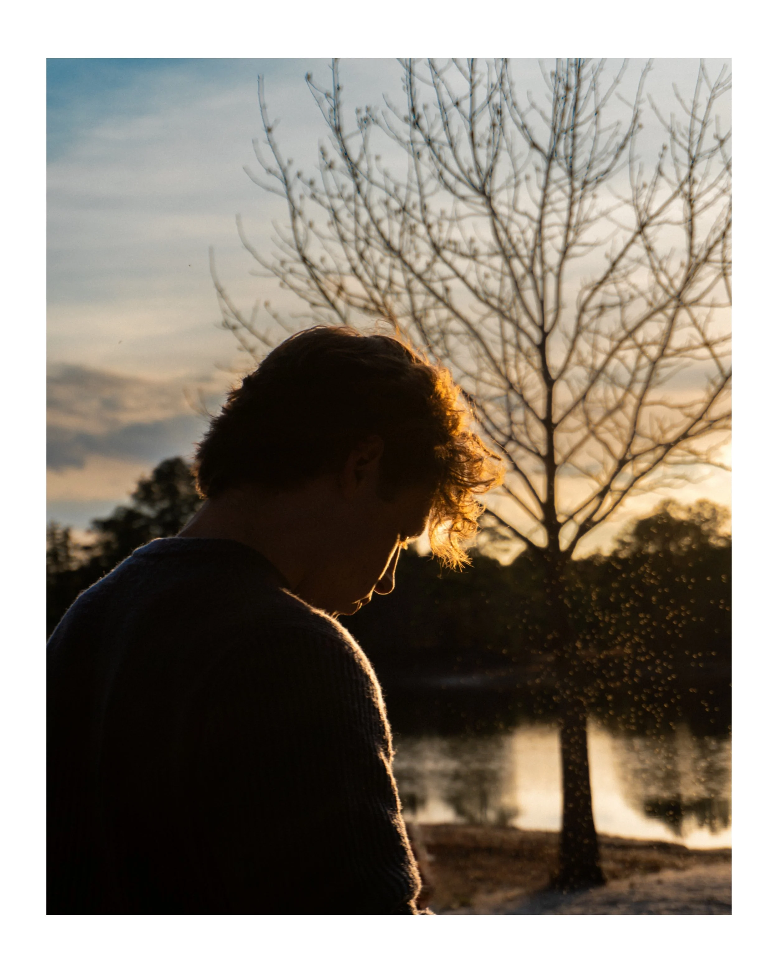 Silhouette of a person with short hair standing outdoors at sunset, with a leafless tree and water in the background.
