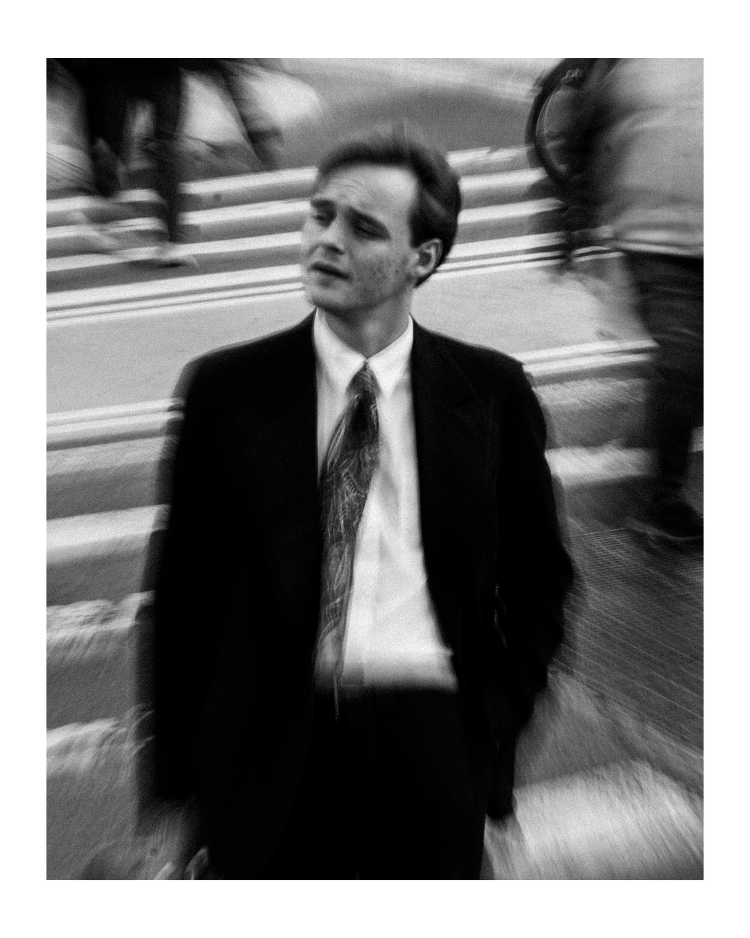 A black and white photo of a man in a suit and tie with a distressed expression, sitting on stairs while people walk by in motion.