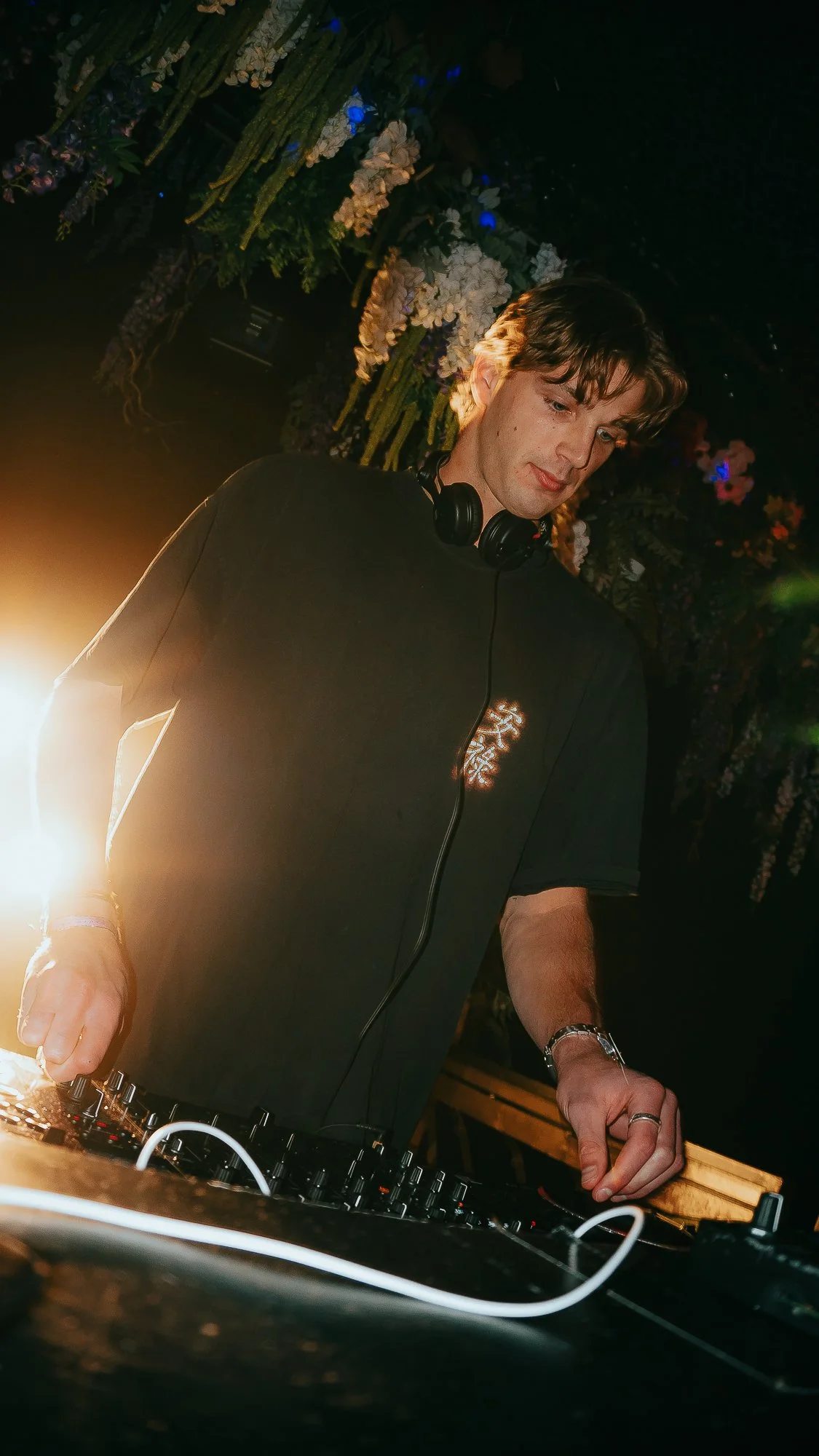 A young male DJ with brown hair and headphones around his neck, wearing a black T-shirt, is performing at a venue decorated with white and purple flowers and green foliage. He is using DJ equipment with cables and turntables in front of him, illuminated by warm lighting.