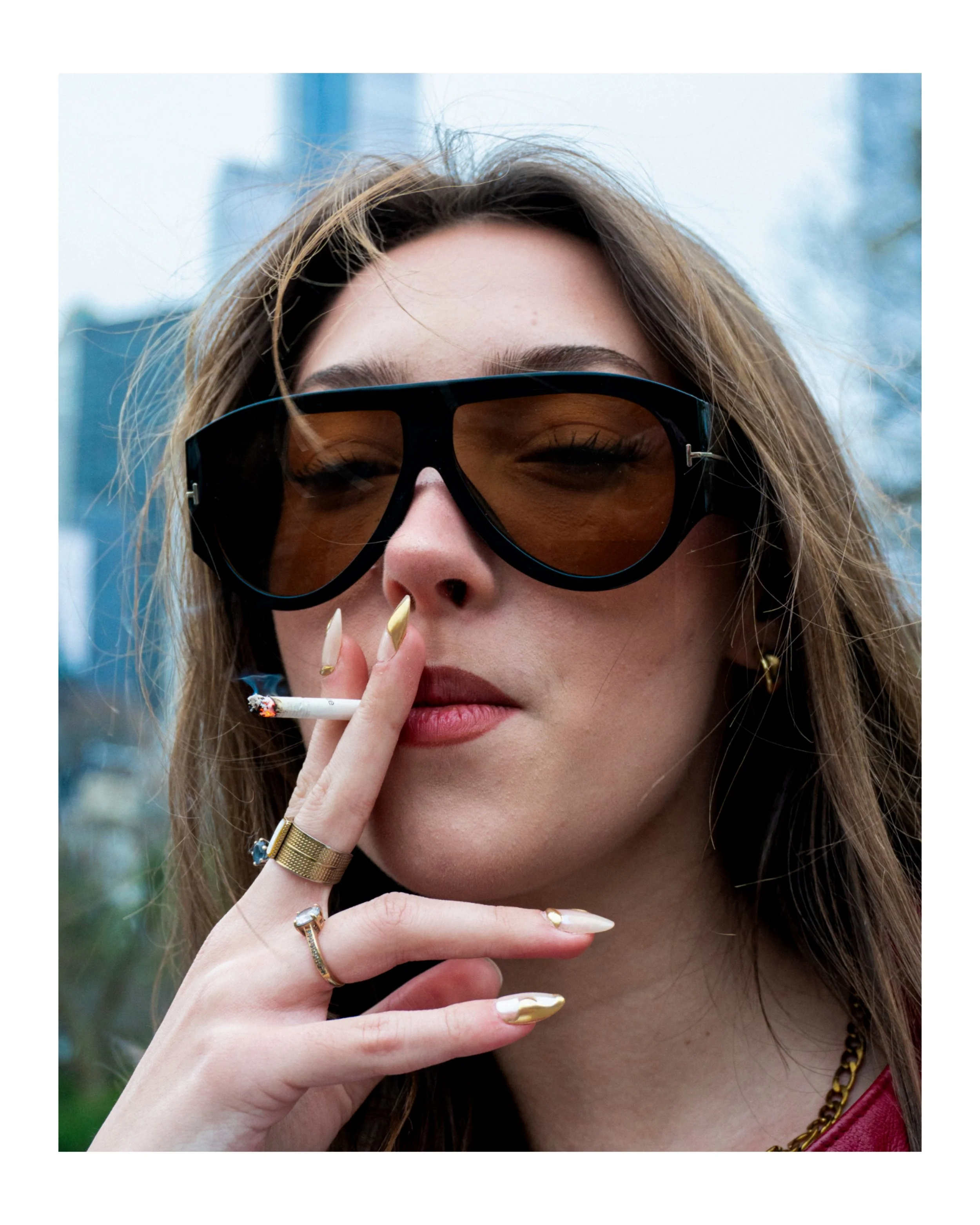 Close-up of a woman with long hair wearing sunglasses, smoking a cigarette, with jewelry including rings, a chain necklace, and earrings, outdoors in a city setting.