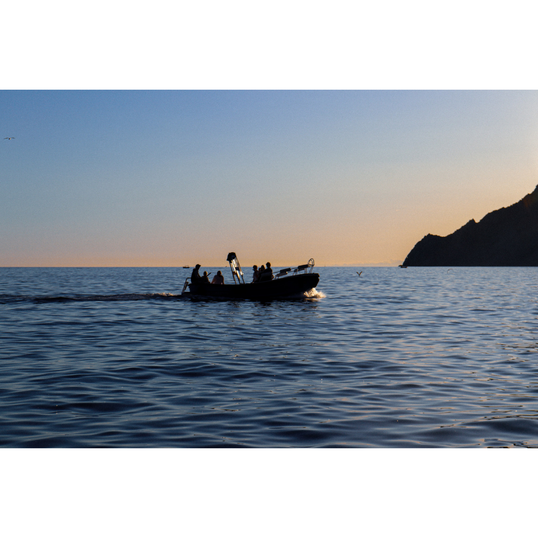 A boat with six people on board is sailing on the water during sunset, with a mountain on the right side and a few small boats in the distance.
