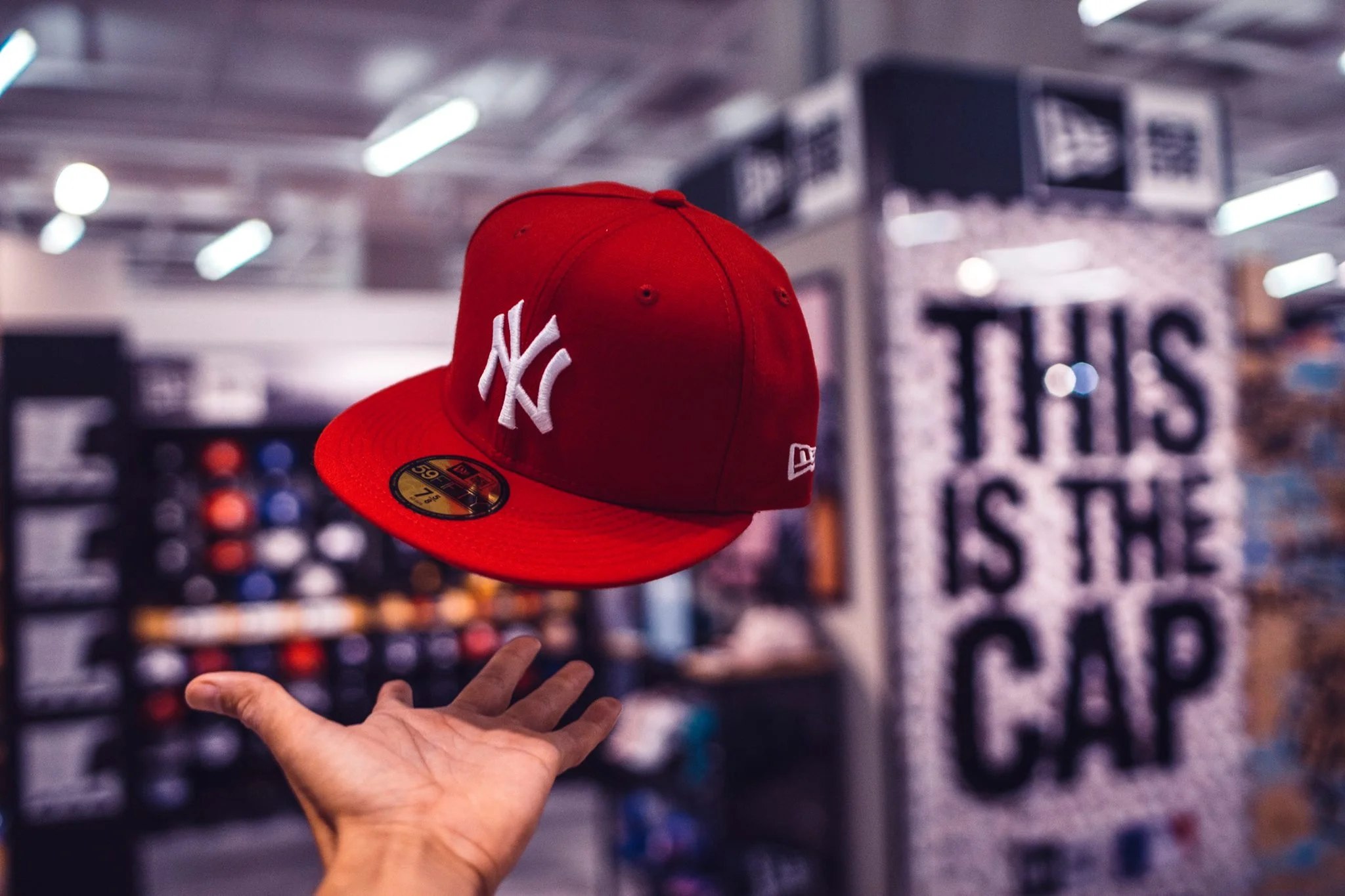 A hand holds a red New York Yankees baseball cap in a store aisle. The background is blurry with shelves displaying various items, and bright overhead lighting.
