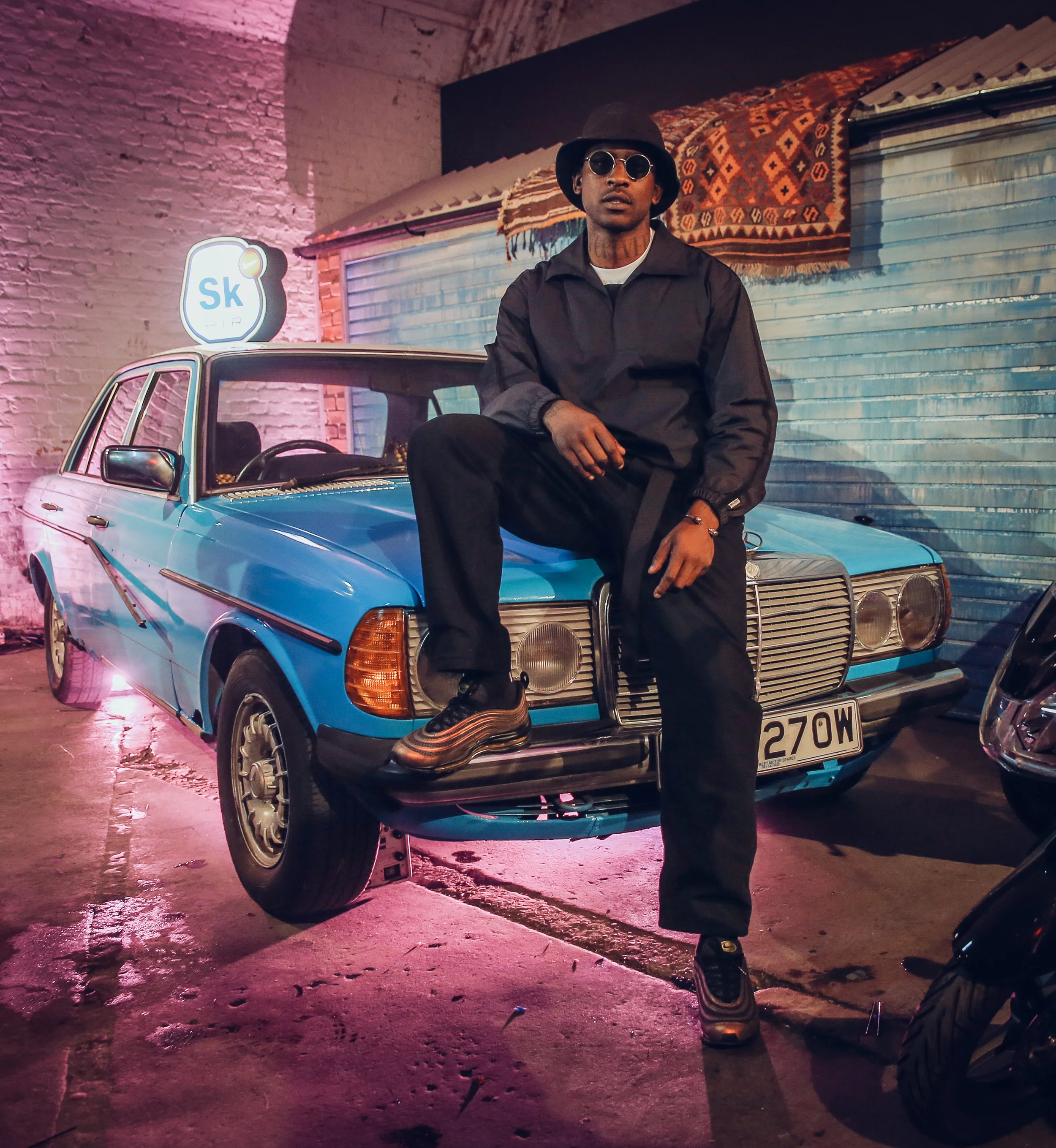 A man wearing sunglasses, a black bucket hat, black jacket, and matching black pants sitting on the hood of a vintage blue car, illuminated with pink and orange neon lights in an urban alley with a brick wall and metal shutter in the background.