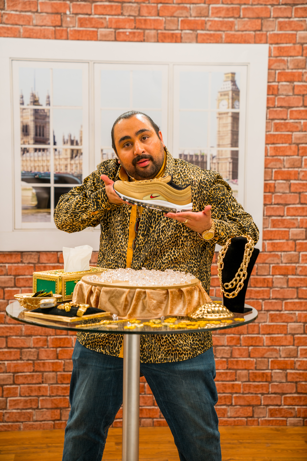 A man with long hair, dark beard, wearing a leopard print jacket, holding a Nike sneaker at a decorated table with gold coins and jewelry, in front of a window with an urban city view.