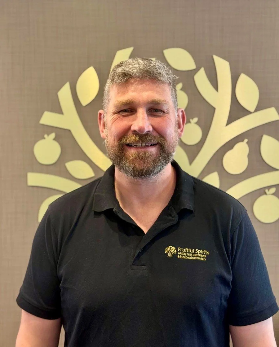Man with graying beard and mustache smiling, wearing a black polo shirt with 'Fruitful Spirits' logo, standing in front of a tree and fruit-themed wall decoration.