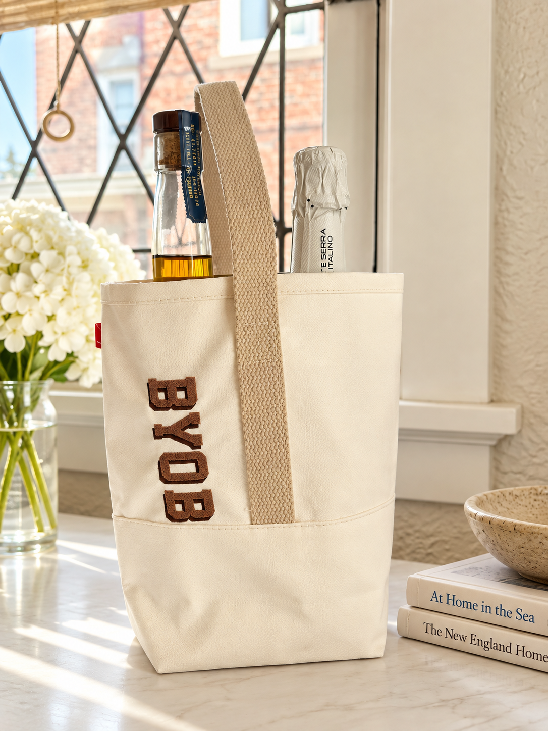 Canvas BYOB wine bag holding two bottles beside white hydrangeas on a sunlit windowsill