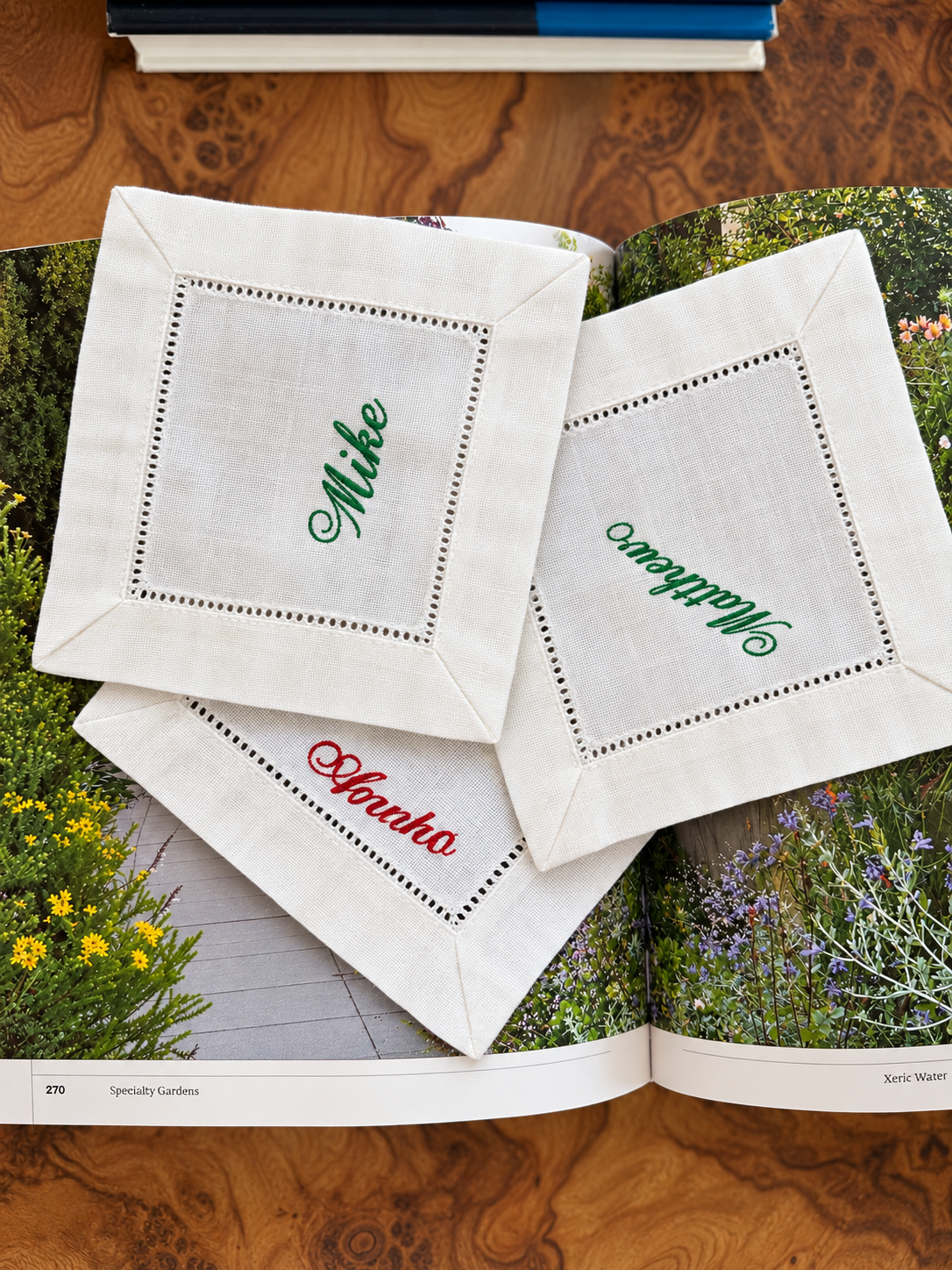 Three linen cocktail napkins embroidered with guest names in green and red script, laid on a garden book