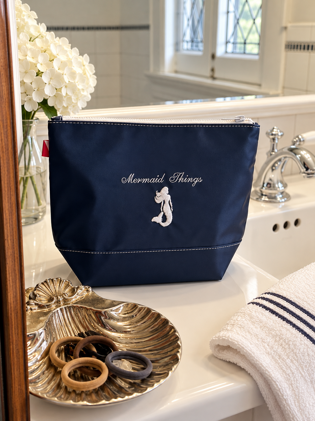 Navy embroidered Mermaid Things pouch on a bathroom counter with white hydrangeas and a striped hand towel