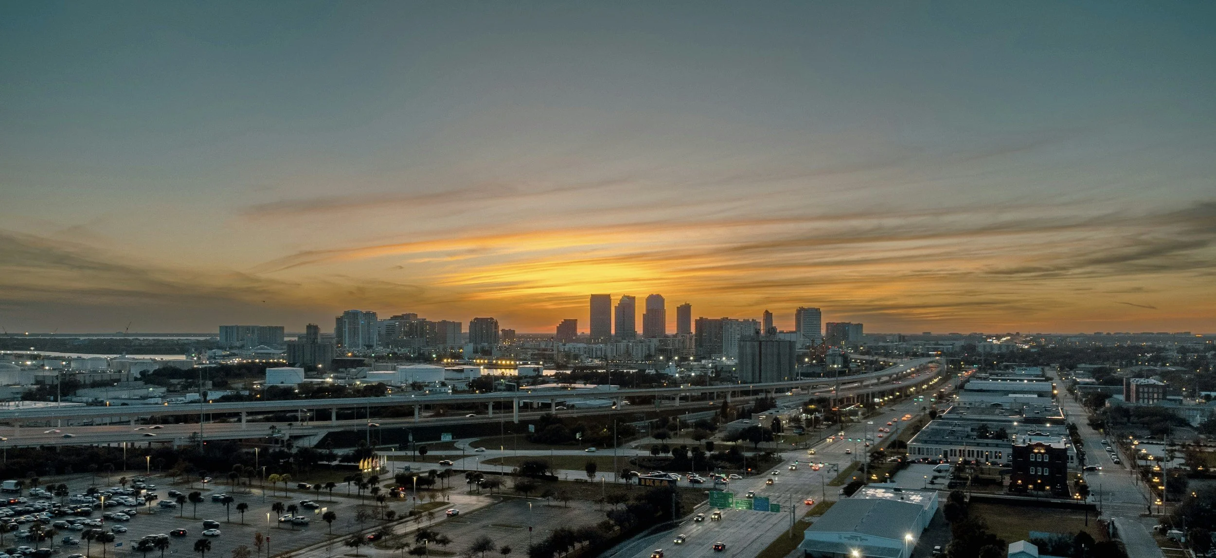 City skyline during sunset with tall buildings, highways, and a parking lot in the foreground.