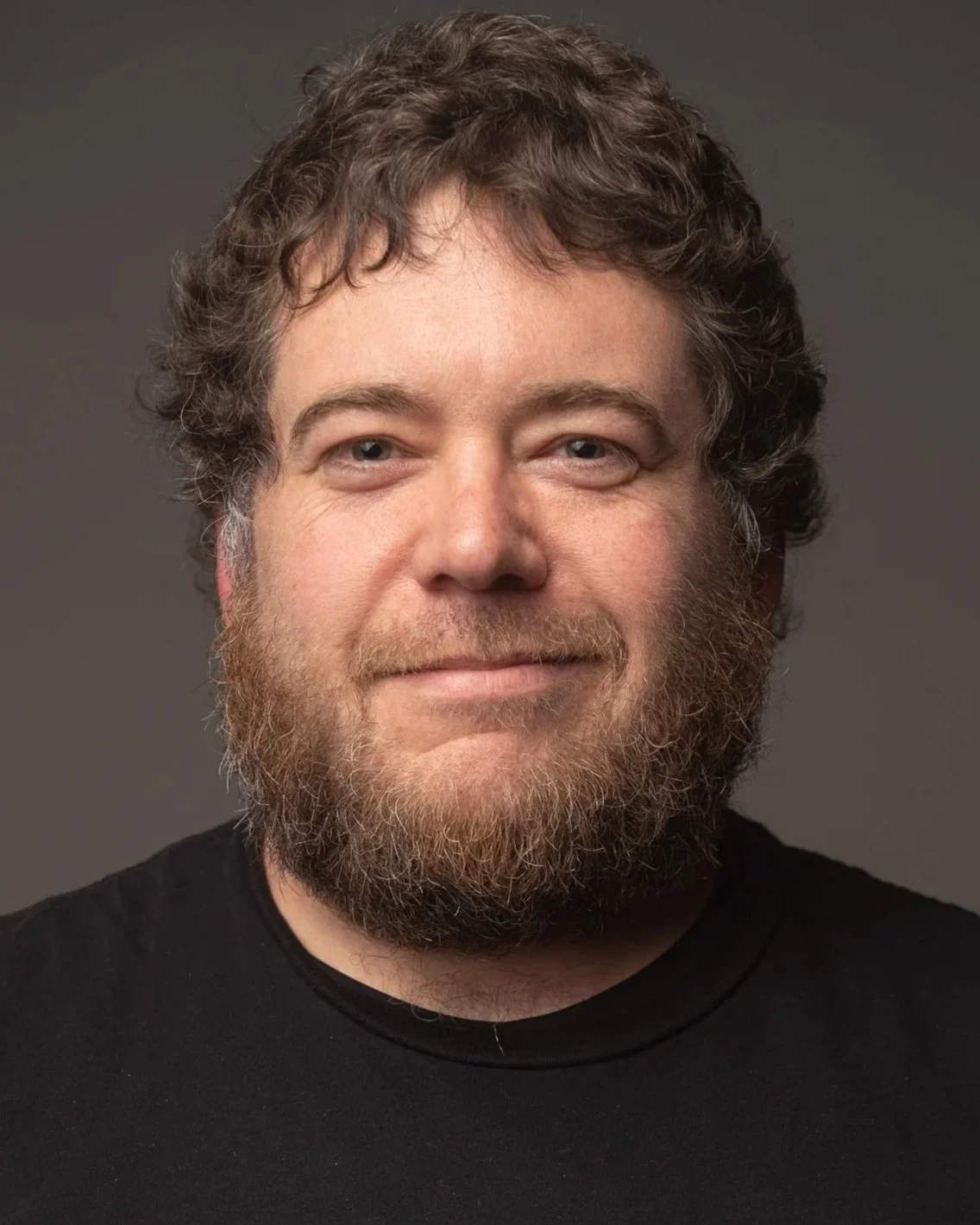 Portrait of a man with curly brown hair and a beard, wearing a black shirt, against a dark gray background.