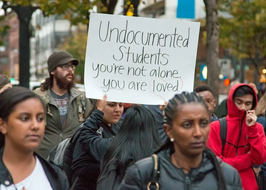 A diverse group of protesters, including women and children, holding a sign that says "Undocumented Students you're not alone you are loved" during a demonstration on a city street.