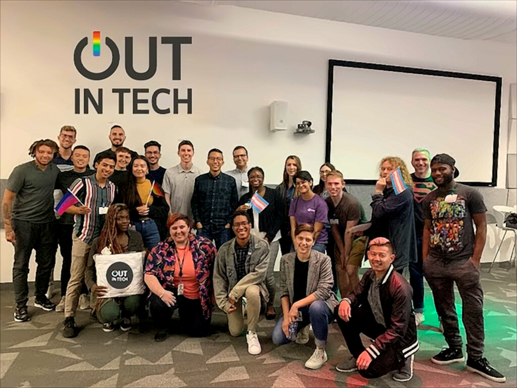 A diverse group of people gathered in front of a wall with a large sign that reads "OUT IN TECH" with the 'O' stylized as a power button. They are smiling, some holding small flags, and appear to be at a tech or community event.
