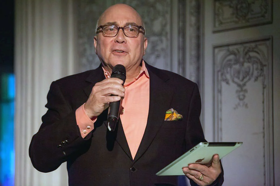 A man in glasses and a dark suit jacket holding a microphone and a tablet appears to be speaking at an event, with elaborate wall decorations in the background.