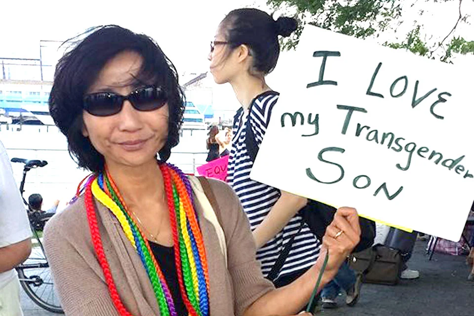 An older woman wearing sunglasses and colorful rainbow beads holding a sign that says 'I Love my Transgender SON'. She is smiling at a public outdoor gathering with other people around.