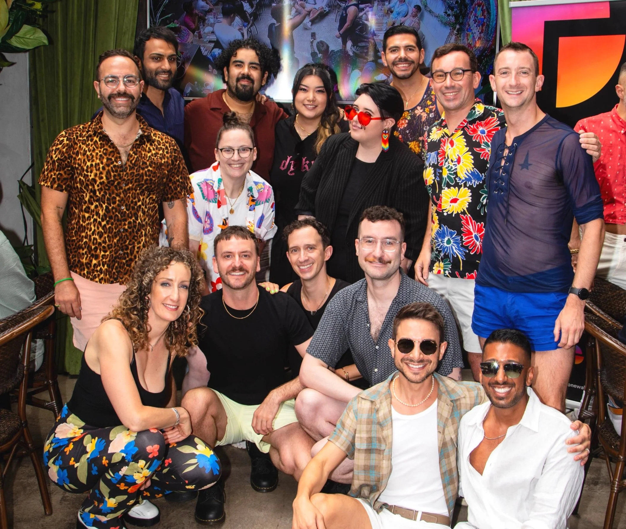 Group of people smiling and posing for a photo indoors, dressed in colorful, casual summer clothing, with a large screen displaying an abstract background behind them.