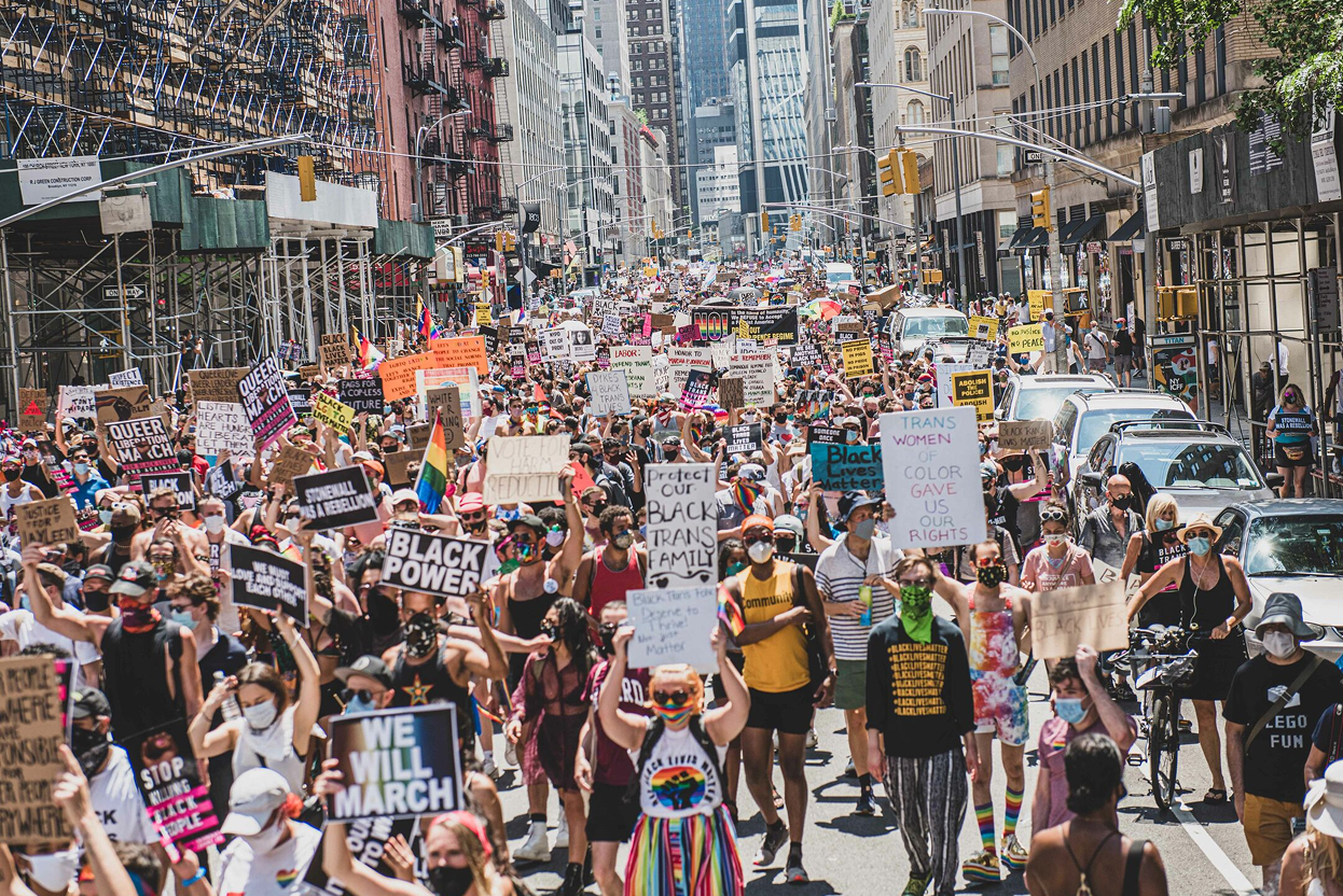 Large crowd of protesters marching on city street holding signs and banners supporting Black Lives Matter, LGBTQ rights, and social justice.