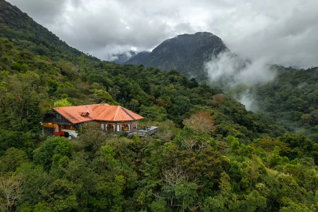 A house with a red roof nestled among lush green trees on a mountain slope, with misty mountains and cloudy sky in the background.