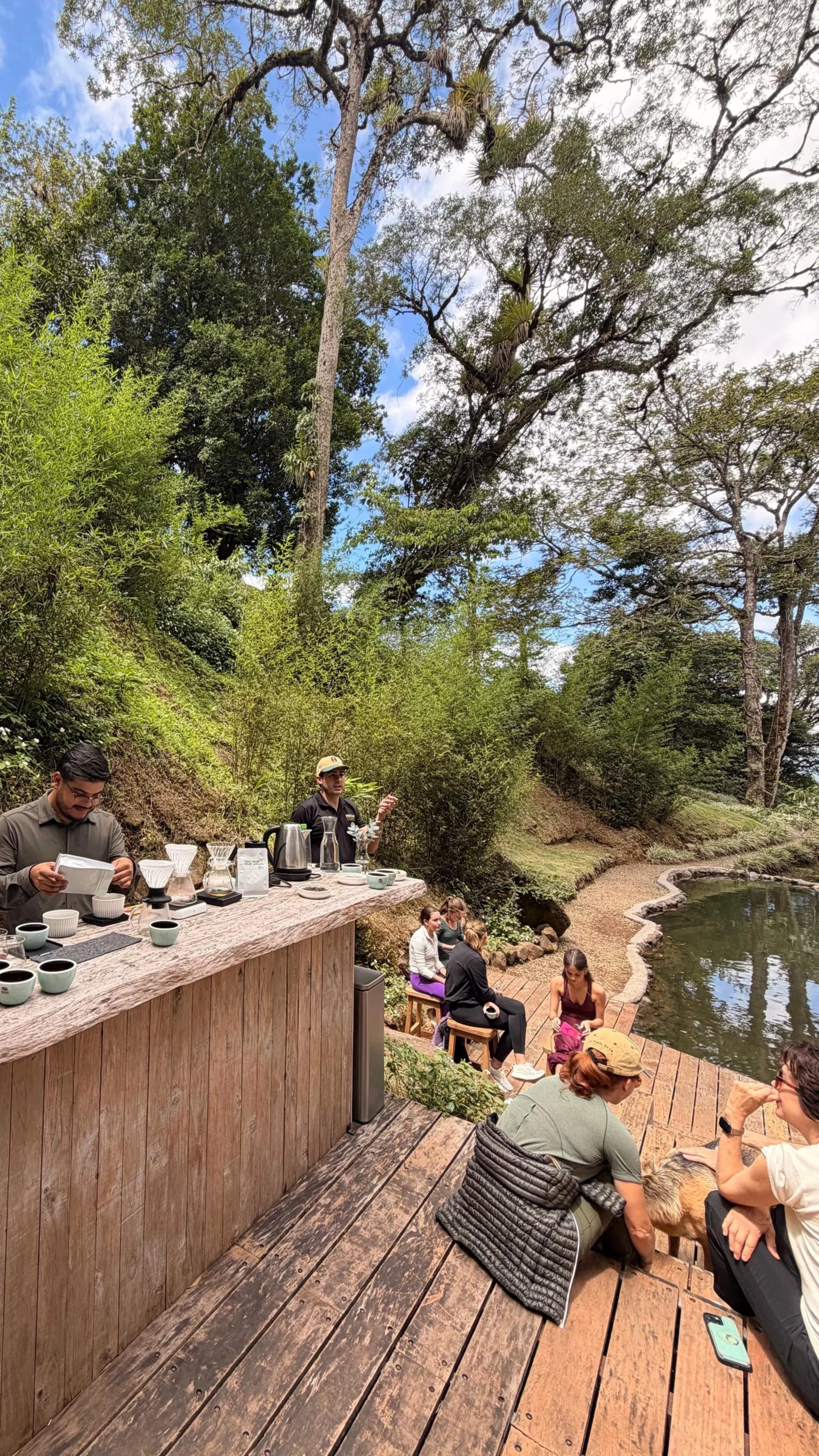 An outdoor gathering with people seated and standing near a riverbank surrounded by trees. A barista is preparing coffee at a wooden counter, while others are sitting on stools and benches, enjoying the natural setting.