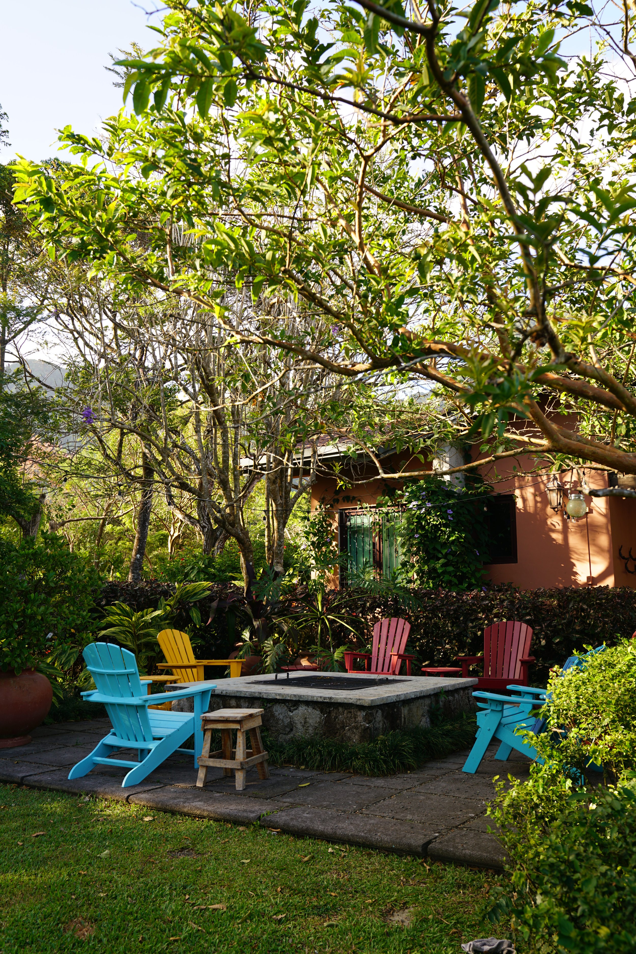 A backyard garden with colorful Adirondack chairs surrounding a fire pit on a stone patio, with lush green plants and trees, and a house with pink walls and lantern lights in the background.