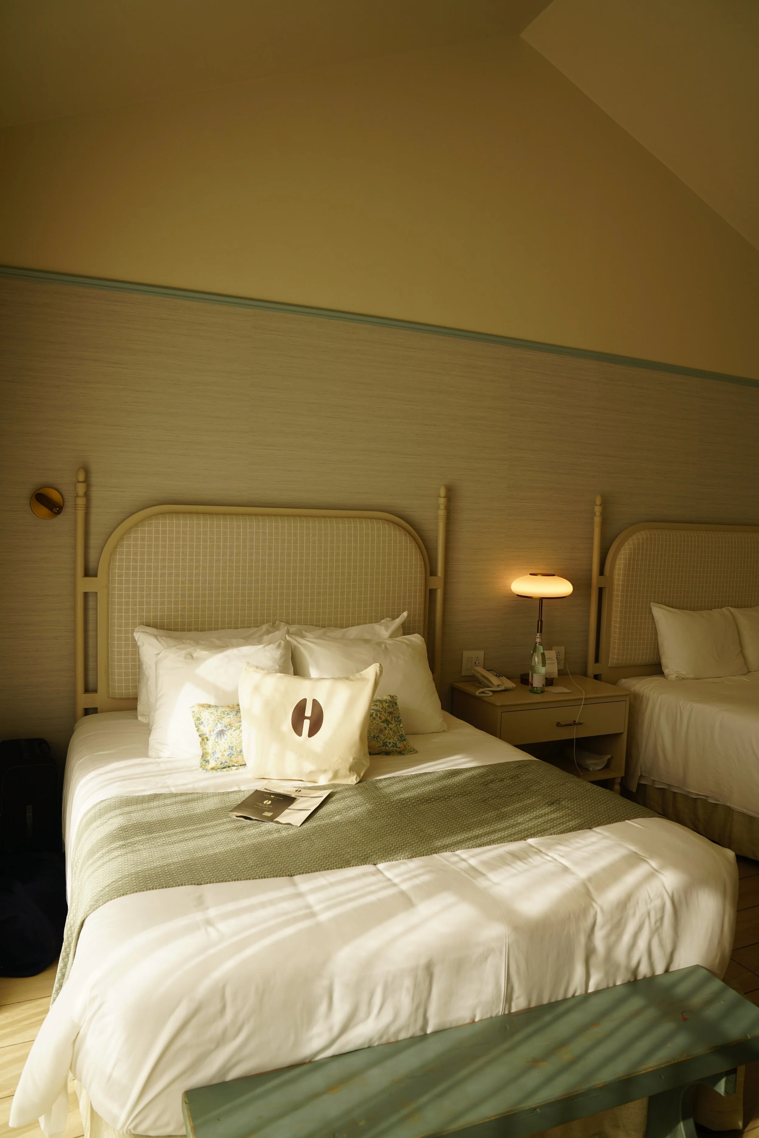 Two neatly made beds with white linens, beige headboards, and a green throw blanket, in a hotel room with beige walls; a nightstand with a lamp, telephone, and water bottle is between the beds.