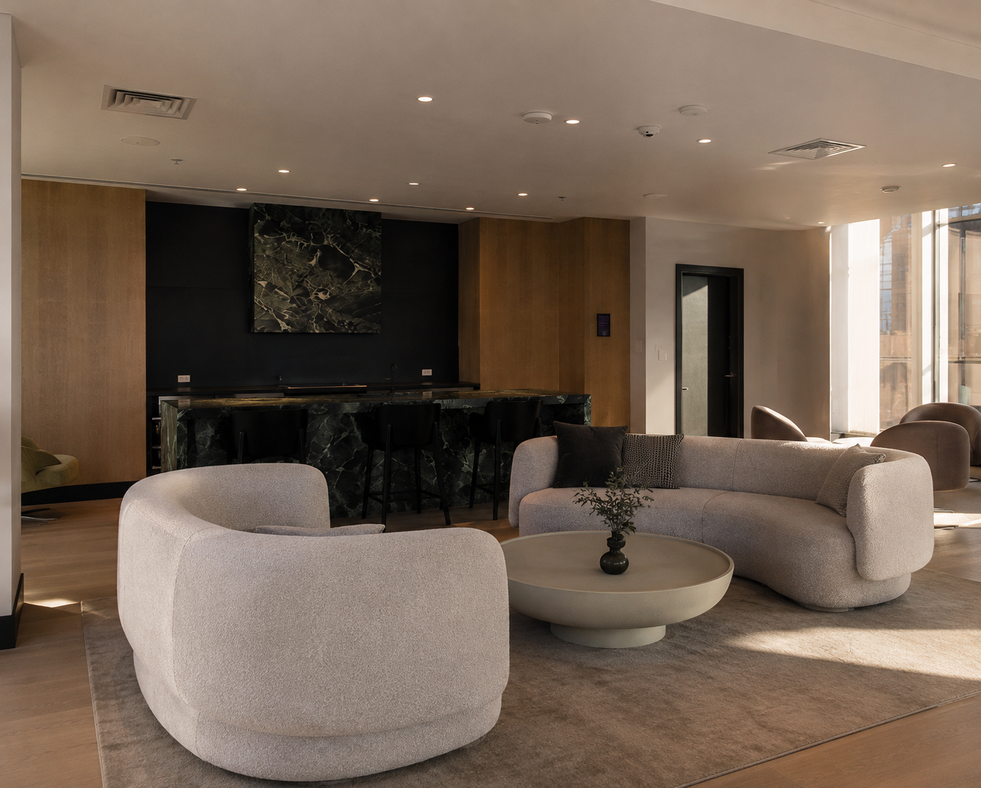 Un salon moderne avec un canapé beige, une table basse ronde avec un vase de plantes, et des fauteuils en peluche. En fond, un comptoir de bar avec des chaises, un mur noir avec une œuvre d'art, et de grandes fenêtres laissant entrer la lumière natur