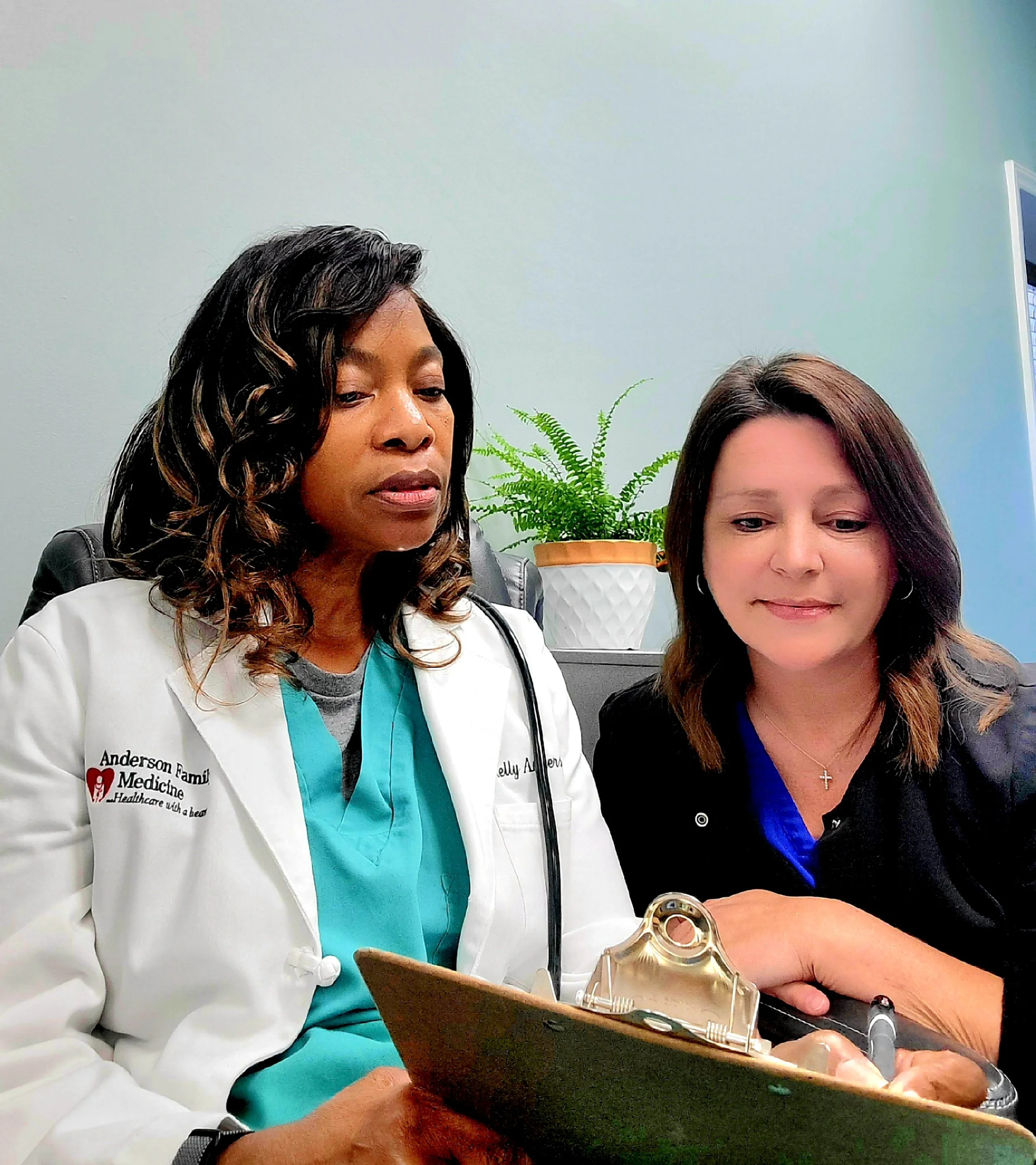 A female healthcare professional in a white coat and teal scrubs filling out paperwork on a clipboard, with another woman sitting nearby, holding a gold award with a clear glass figure on top. They are in a medical office with a green potted plant on a table behind them and a light blue wall with a window.