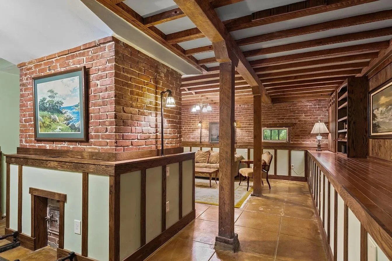Living area with brick walls, wooden beams, and a built-in wooden shelf with a lamp, with a small seating area in the background.