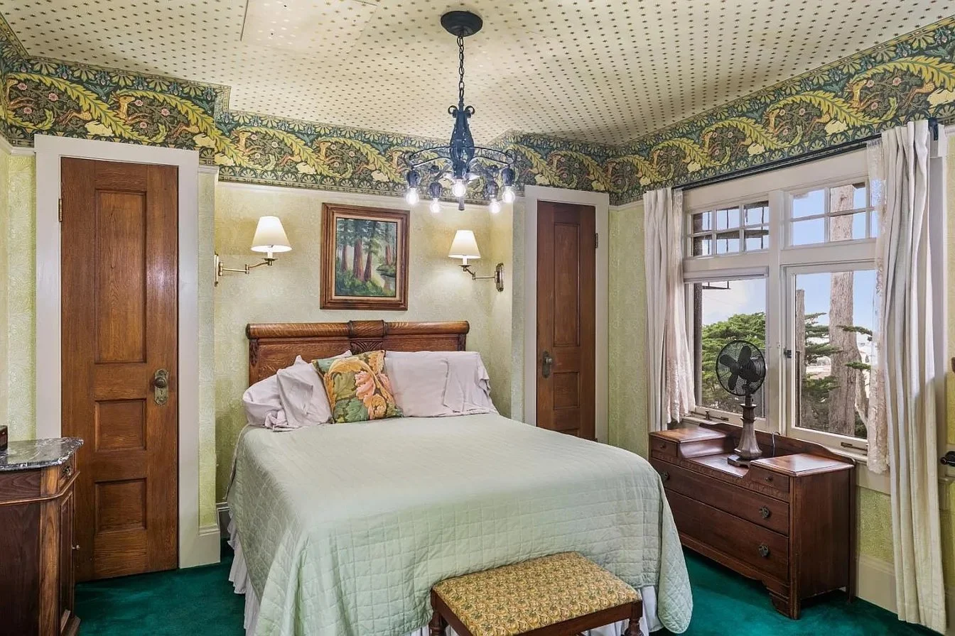 A cozy bedroom with a bed, wooden furniture, a painting, and natural light from a window.