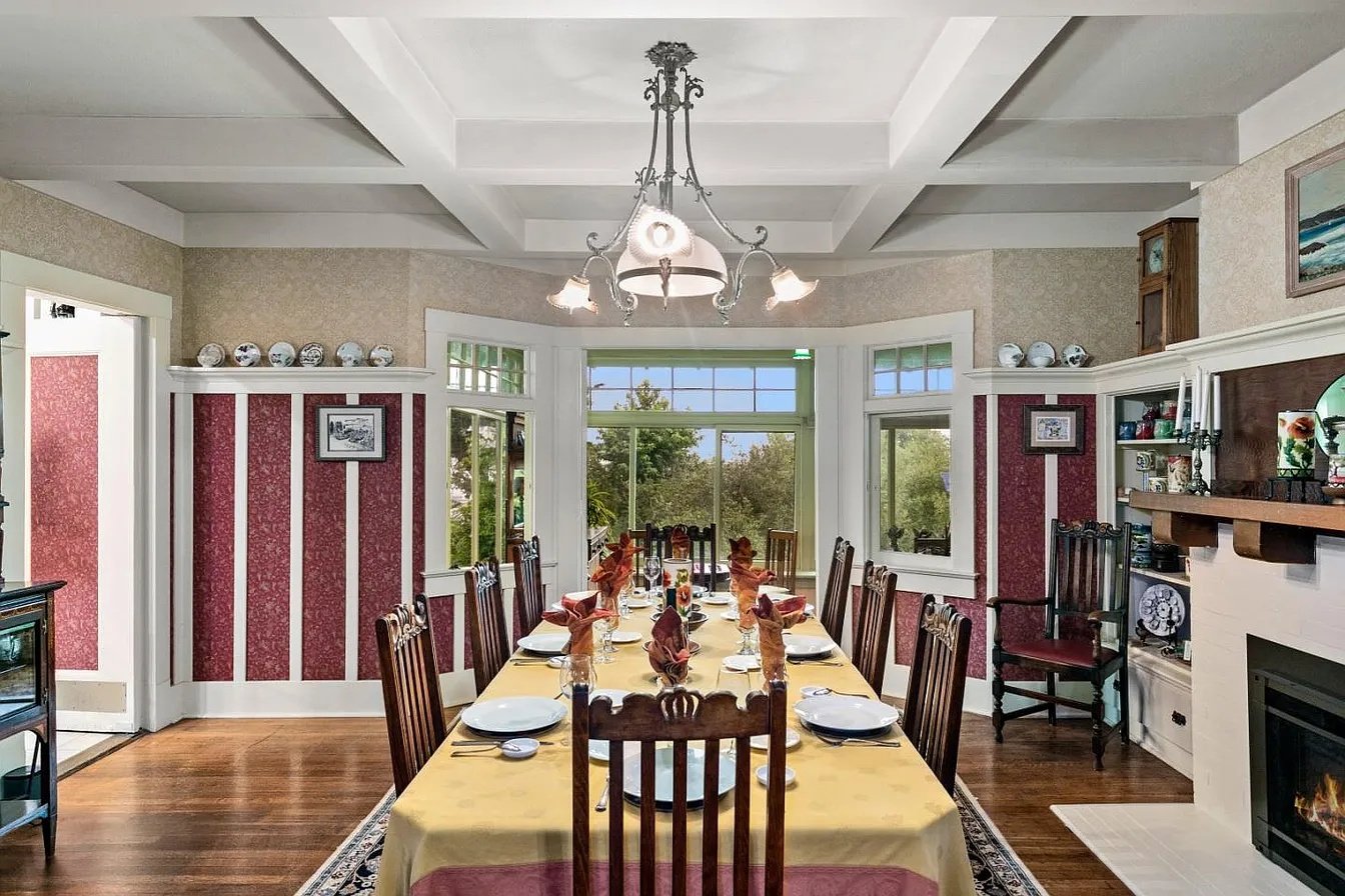 Dining room with a long table set for a meal, surrounded by wooden chairs, a chandelier overhead, and a large window showing trees outside.