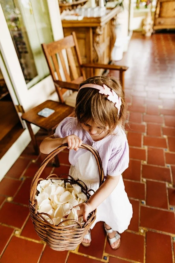 A young girl with a pink headband holding a basket filled with white flower petals on a terracotta-tiled porch.