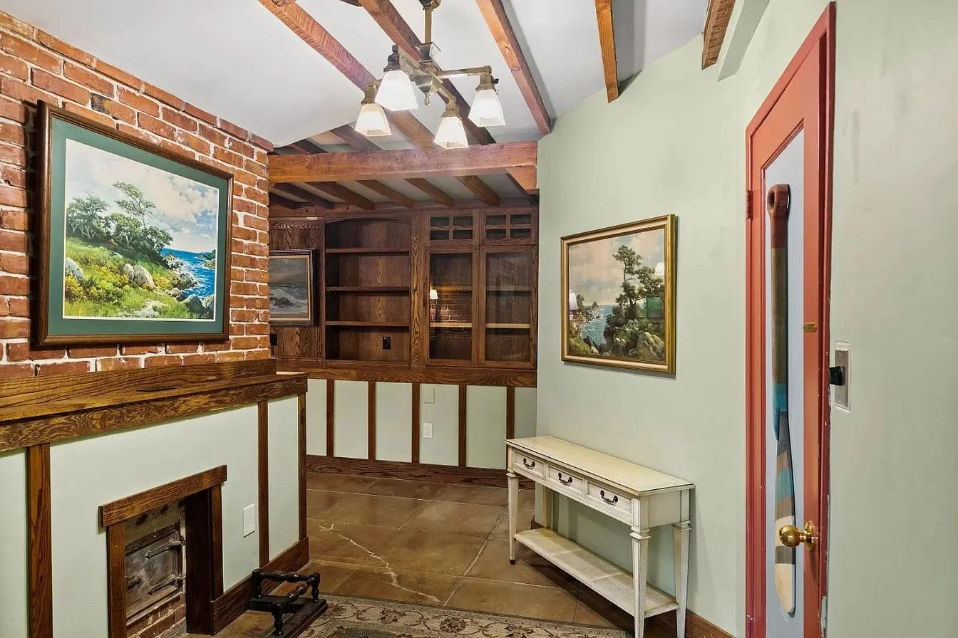 Interior of a house with painted and brick walls, wood ceiling beams, a white table with drawers, framed landscape paintings, and a ceiling light fixture with five lights.