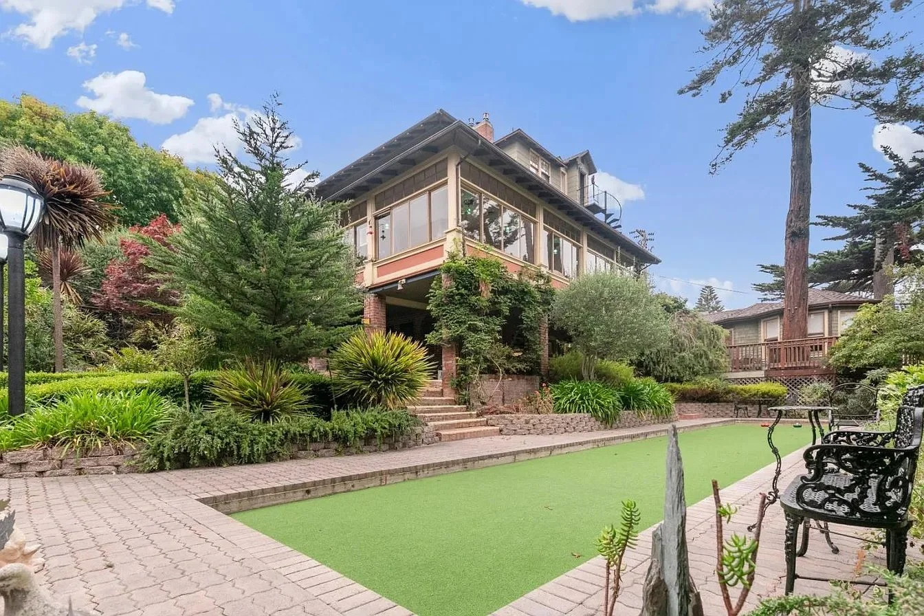 A backyard with a brick patio, green artificial turf, black wrought iron chairs, lush garden plants, and a large house with a screened porch on a sunny day.